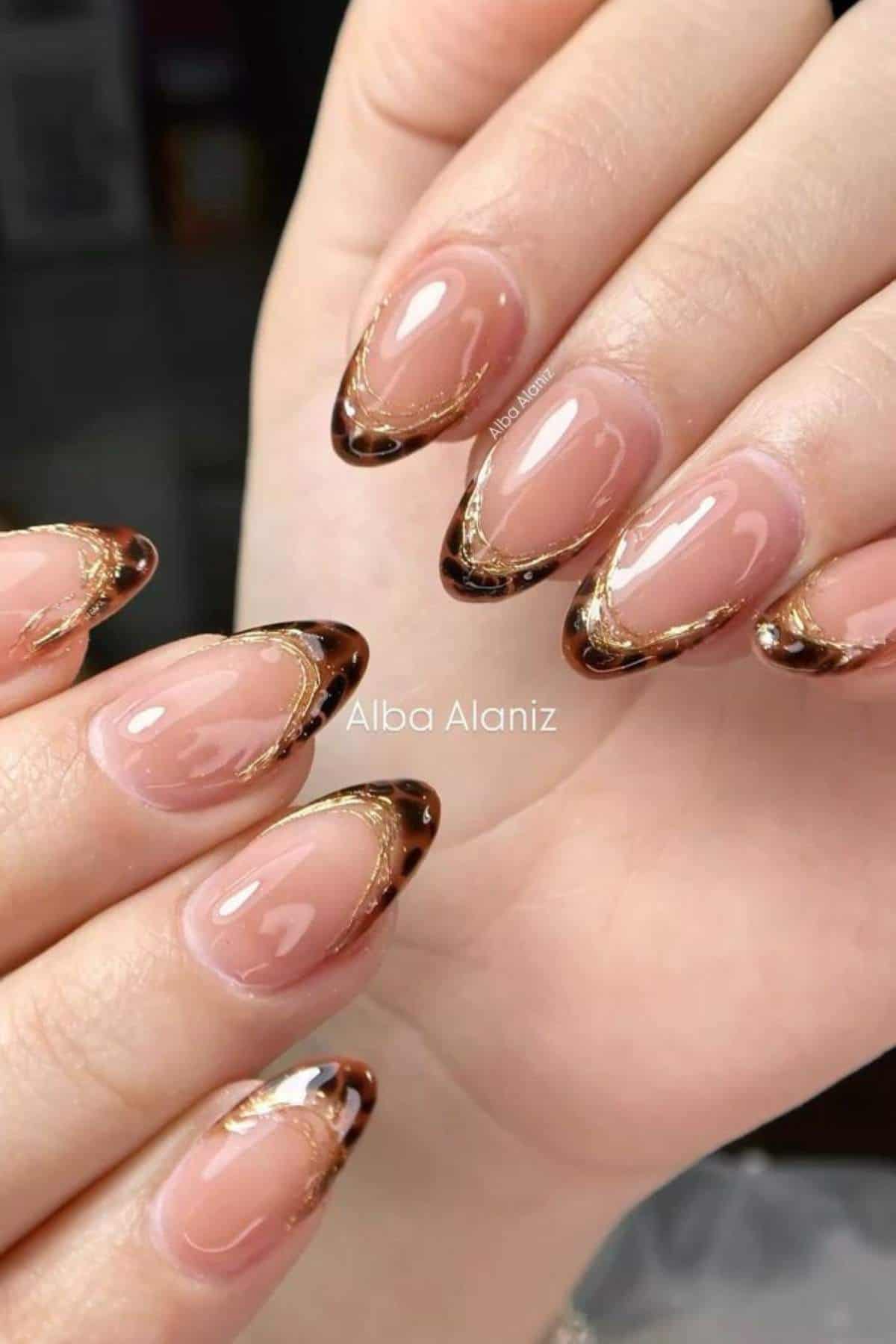 Hands with almond-shaped nails featuring a nude base and brown French tips with gold accents.
