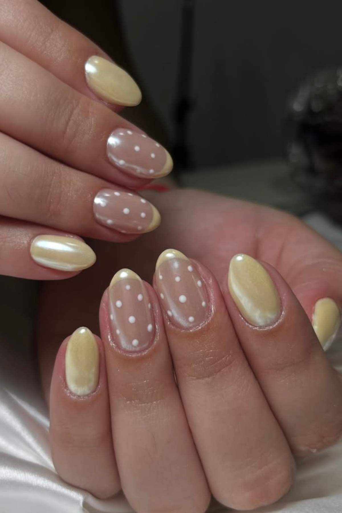 Close-up of a hand with almond-shaped nails featuring pale yellow polish and accent nails with sheer polish and white polka dots.