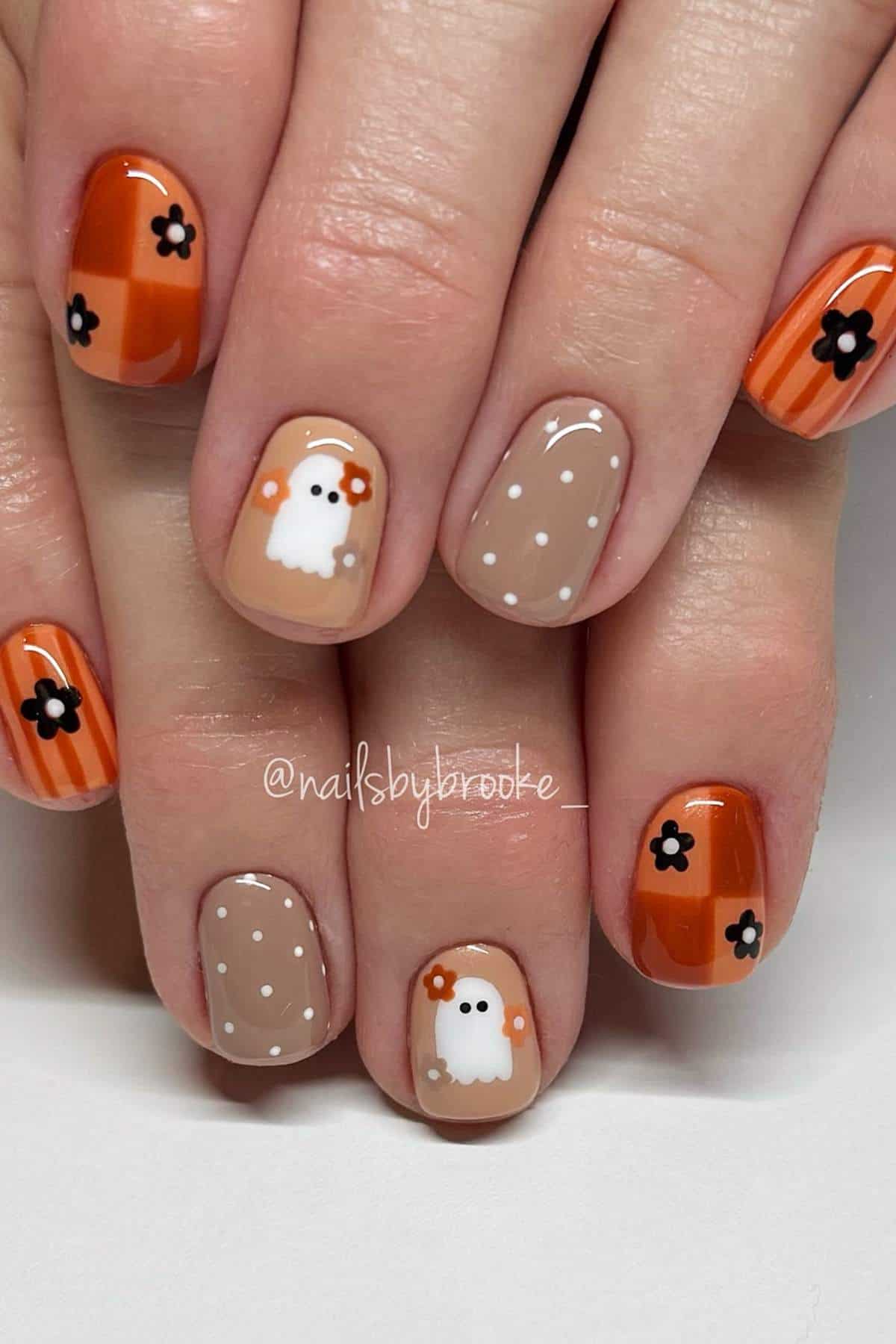 Hands with fall-themed nail art featuring orange stripes, black flowers, white polka dots, and cartoon ghost designs on a tan and orange background.