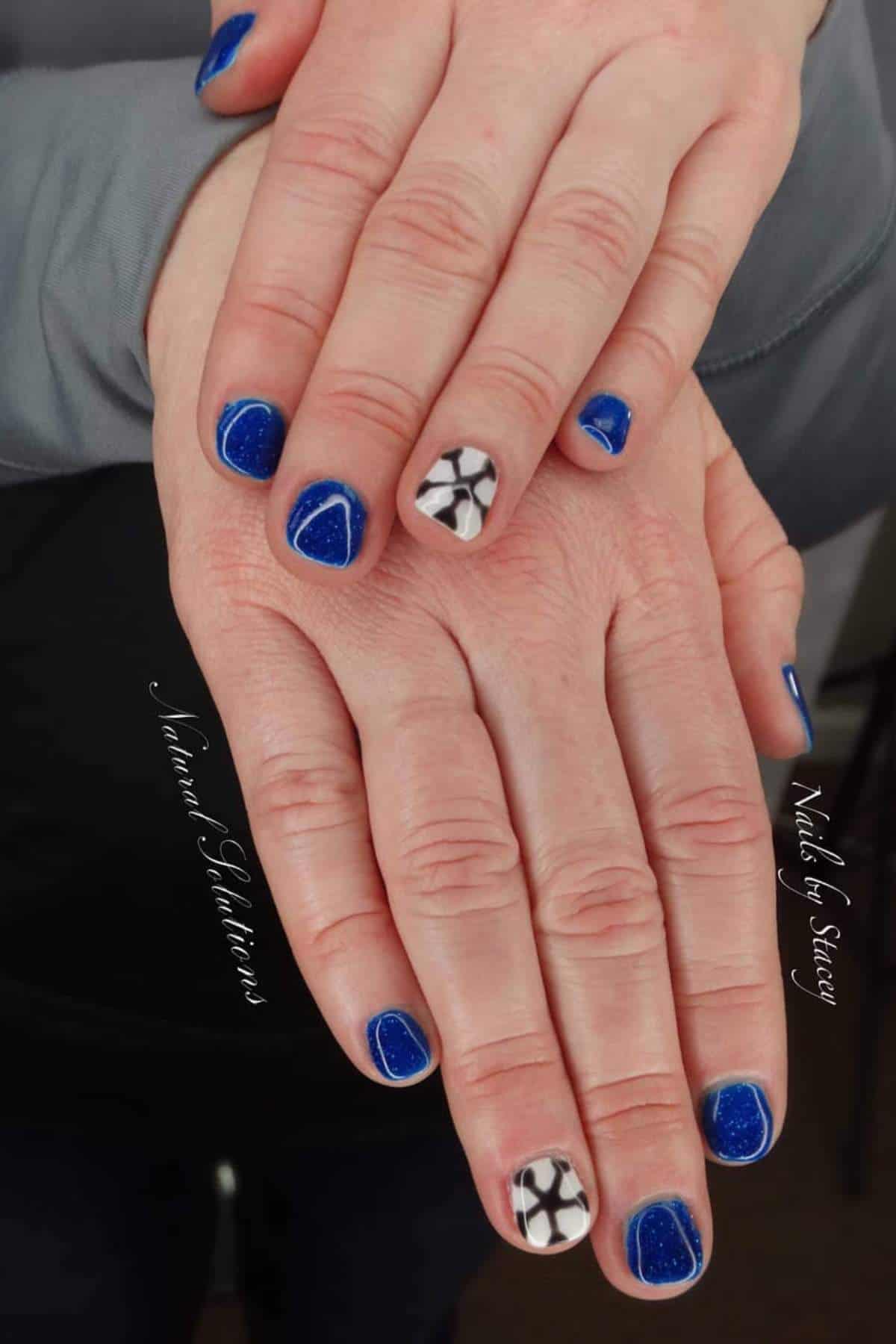 A person’s hands display short, blue-glittered nails with triangular tips; one nail on each hand features a black and white geometric design.