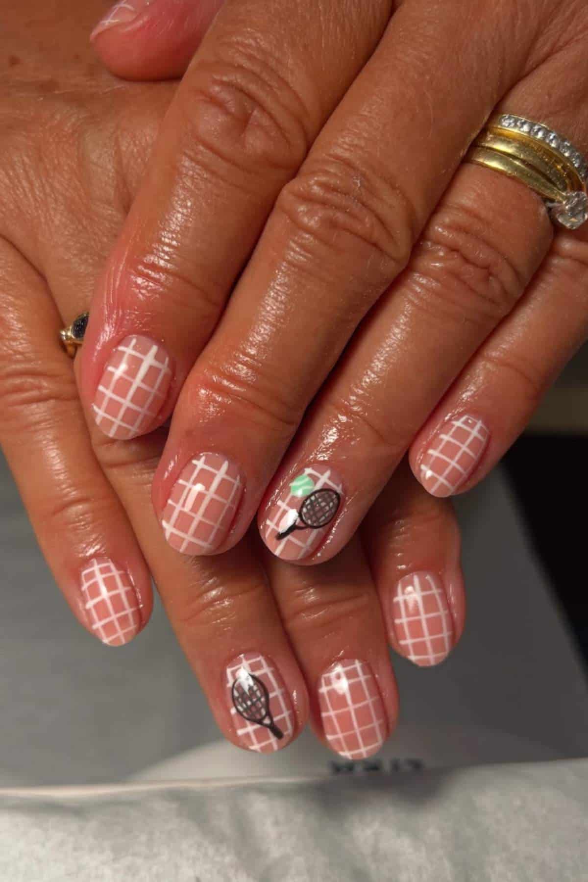 Hands with manicured nails featuring a white grid pattern on a nude base and tiny tennis-themed designs on two accent nails.