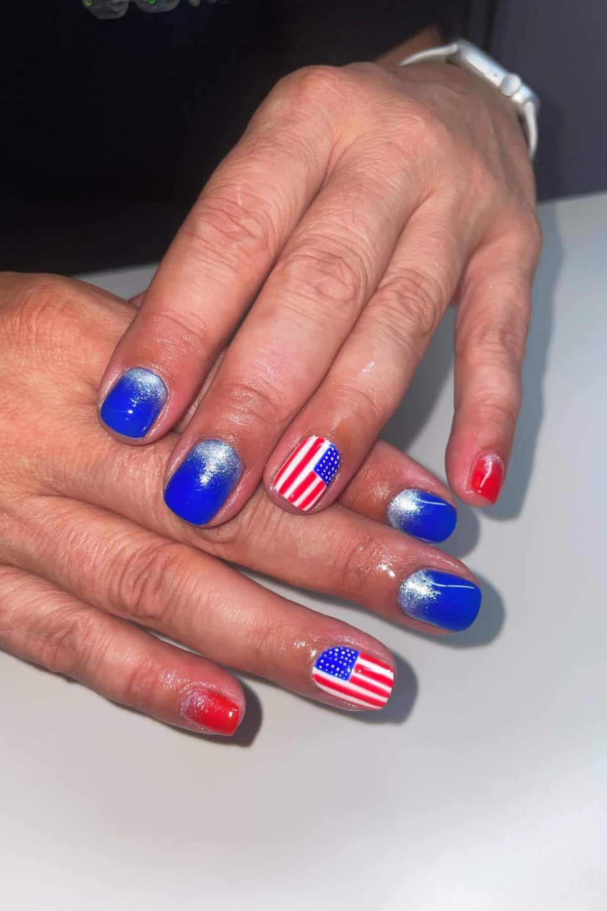 A hand with manicured nails painted in blue, red, and silver, with two nails featuring an American flag design.