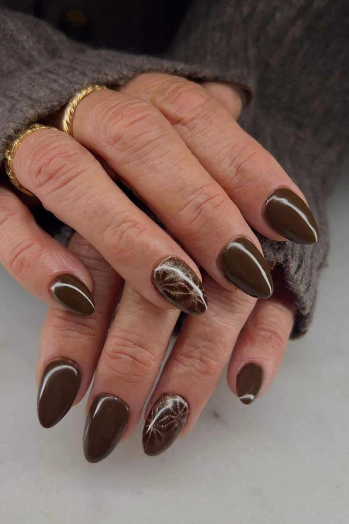 Close-up of hands with almond-shaped brown nails; one accent nail on each hand features a marbled white and brown design.