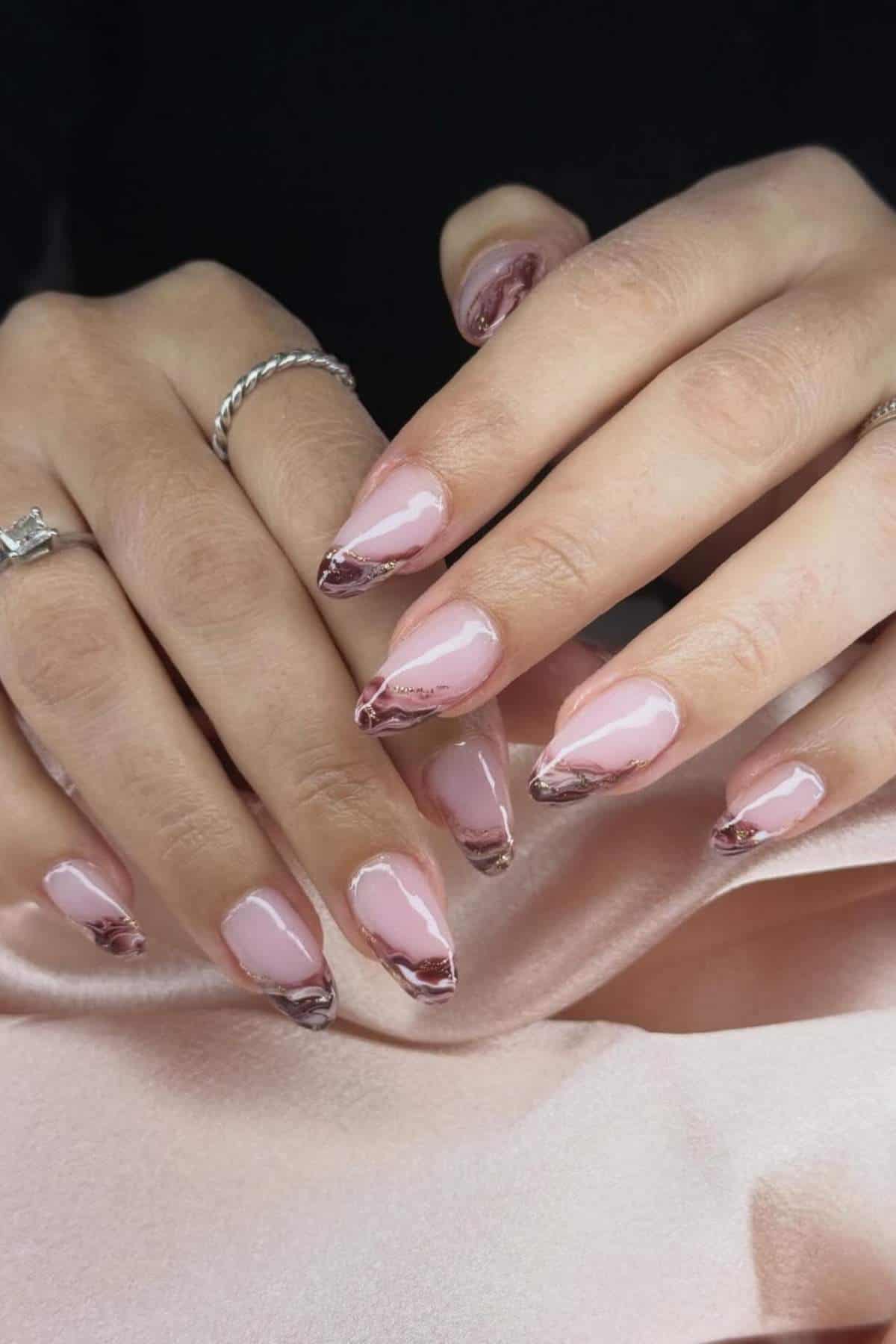 Two hands with almond-shaped nails featuring a pink base and metallic maroon tips. The person is wearing a silver ring and a twisted band ring.