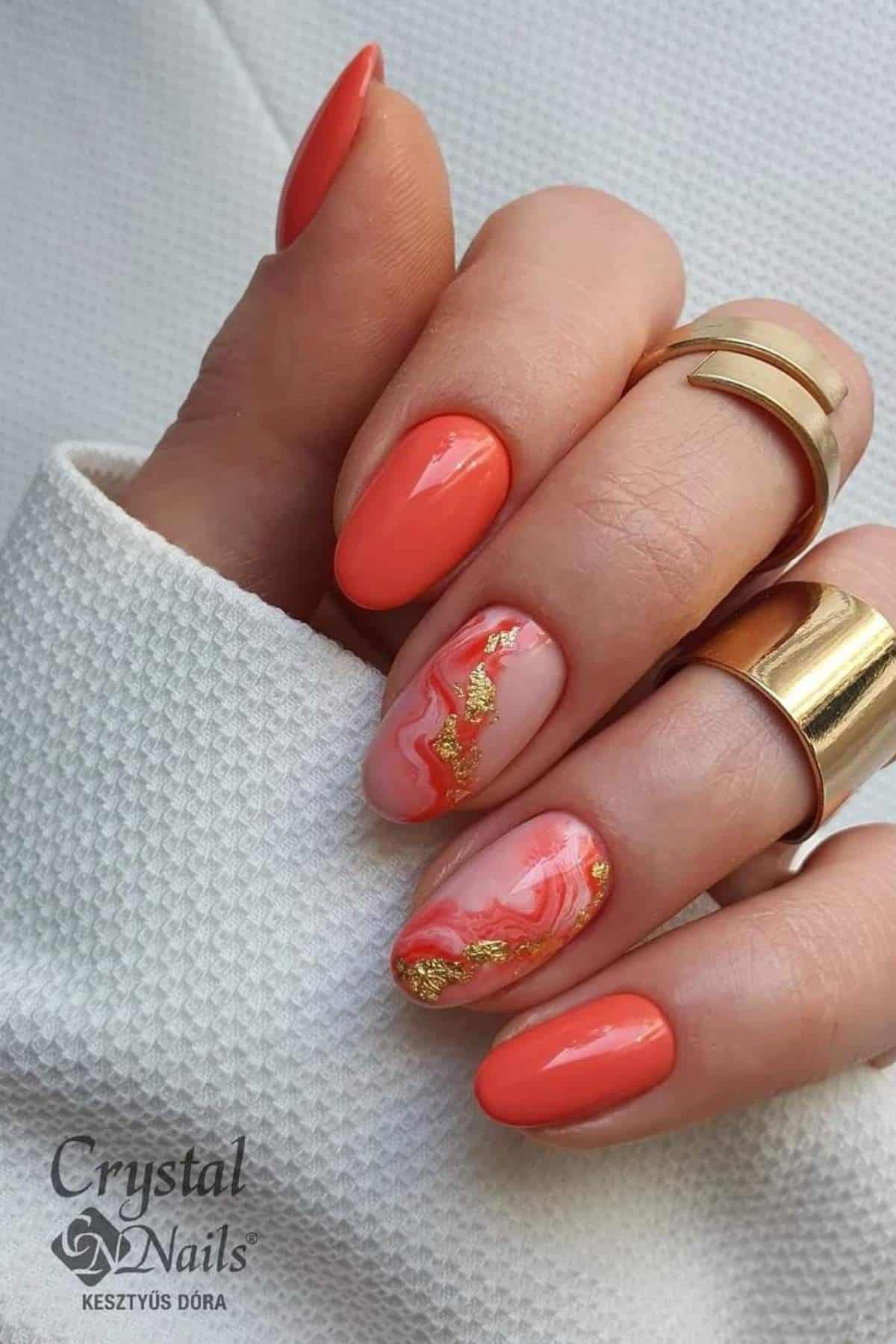 A hand with coral-colored almond-shaped nails, one accent nail featuring gold foil and a pink marble design. The person is wearing gold rings and a white textured sleeve.