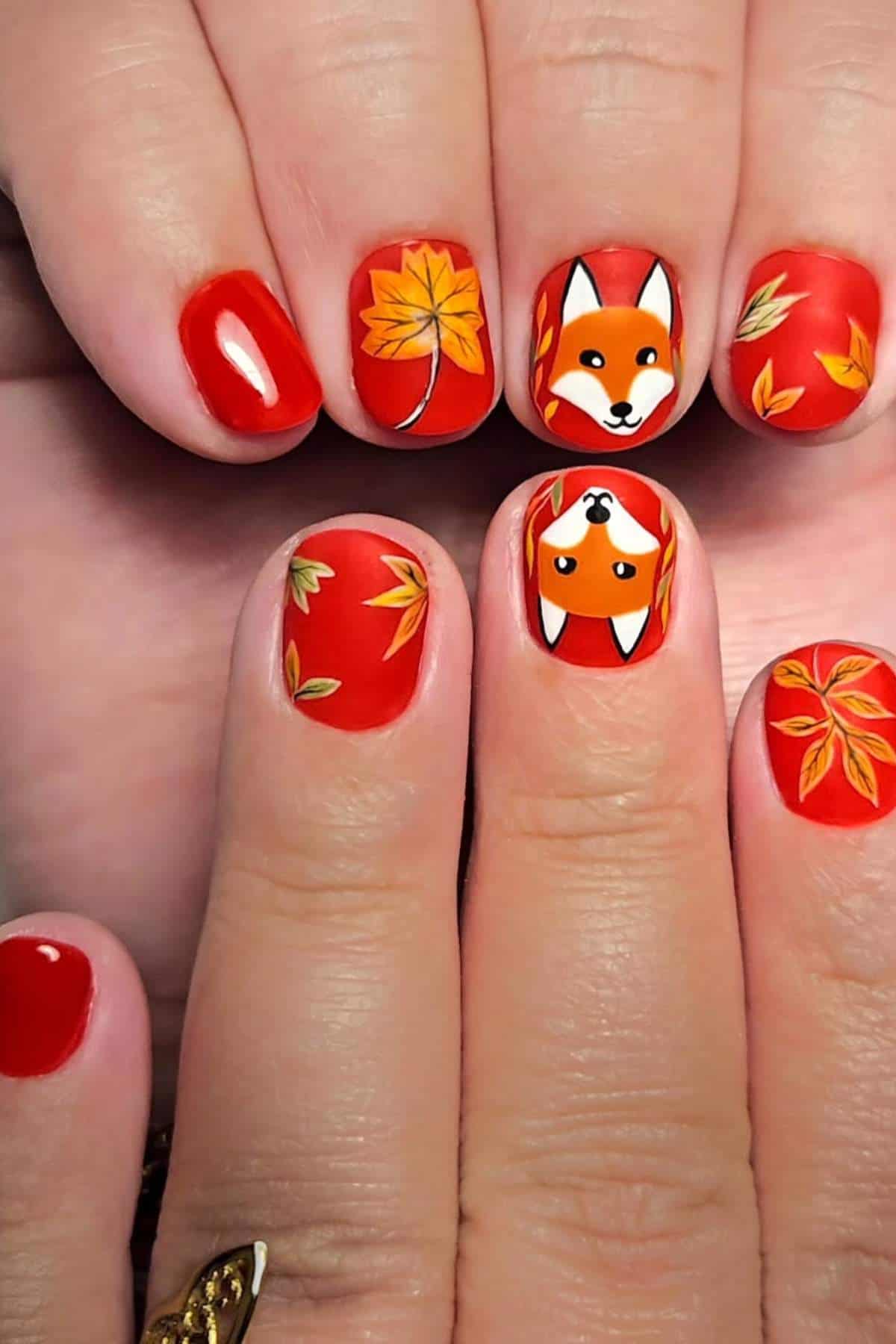 Fingernails painted red with designs of orange autumn leaves and cartoon fox faces on the middle and ring nails.