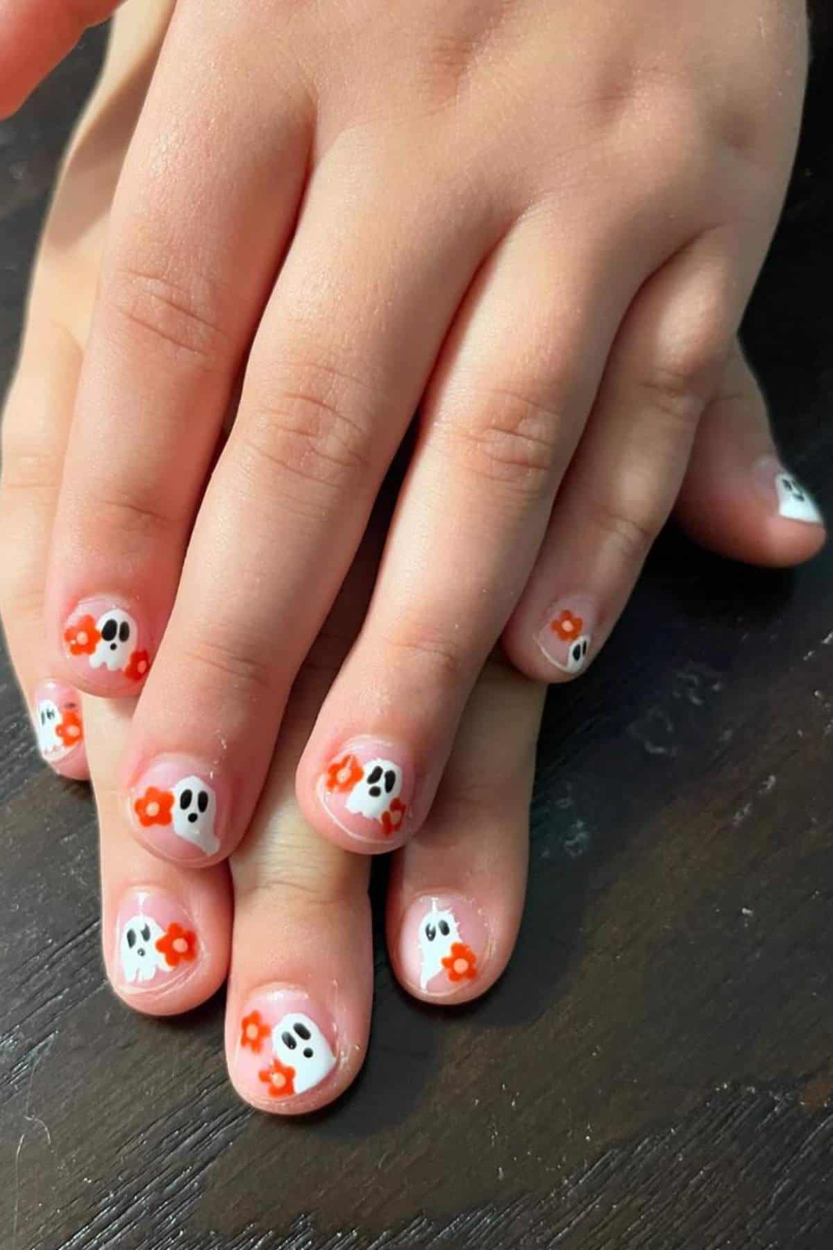 Child’s fingernails with a manicure featuring small ghost faces and orange flowers painted on a clear base.