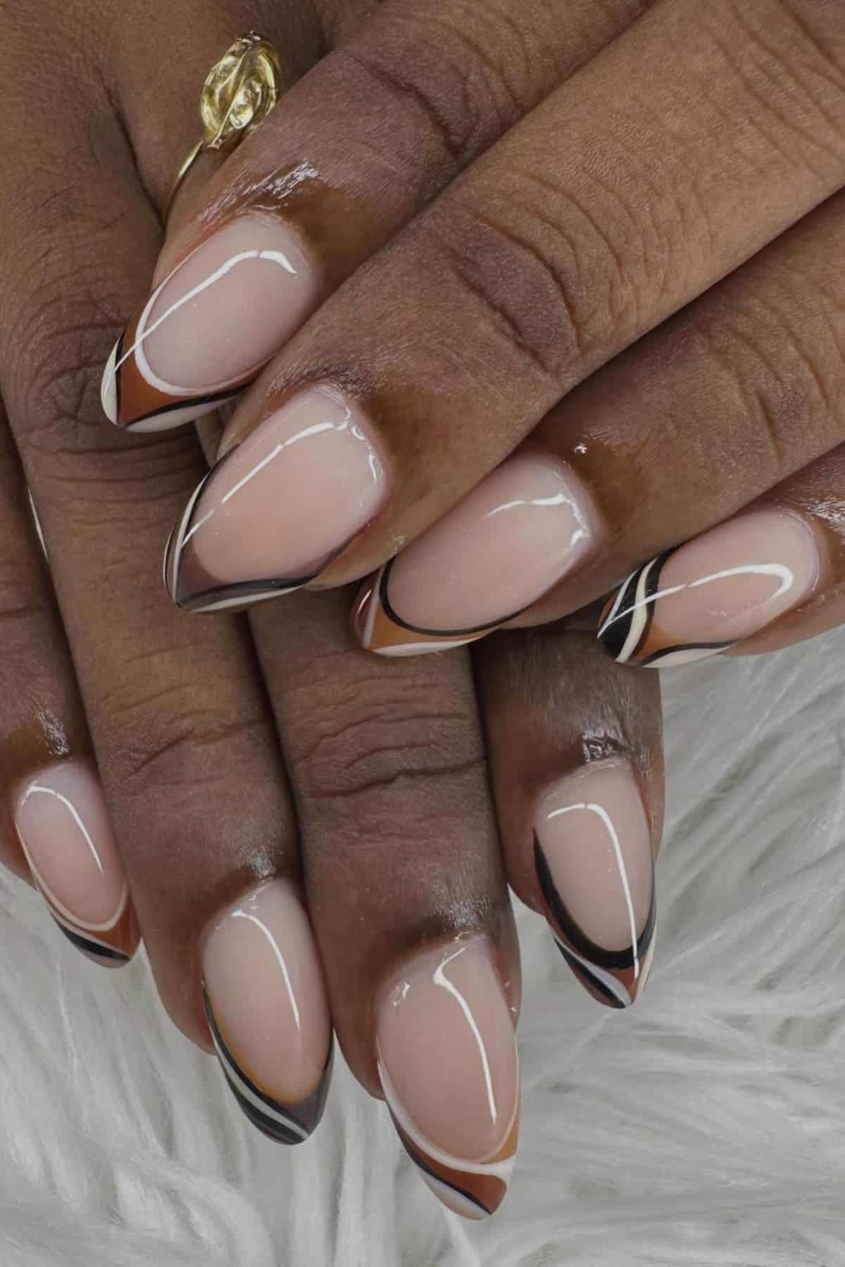 Hands with almond-shaped nails featuring a nude base and black, brown, and white abstract line designs on the tips, resting on a white textured surface.