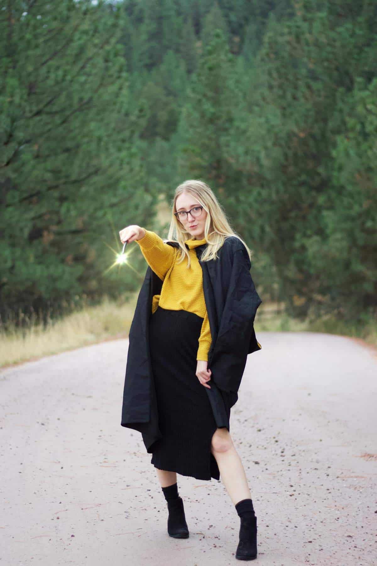 A person in a yellow sweater and black coat stands on a forest road, holding out their hand with a bright light effect at their fingertip.