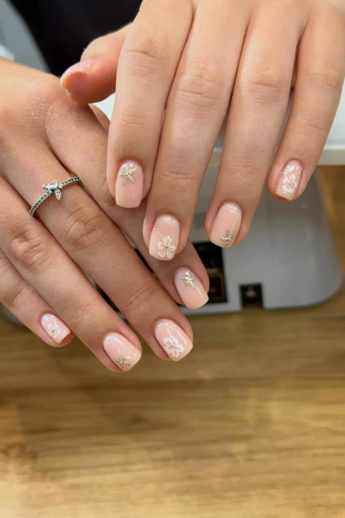 A pair of hands with short, light pink nails featuring white floral designs and small gold star embellishments, resting on a wooden surface.