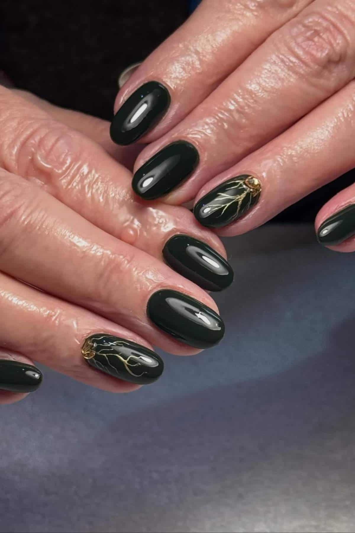 Hands with short, oval-shaped nails painted dark green; two nails feature gold branch-like designs and small gold embellishments near the cuticle.