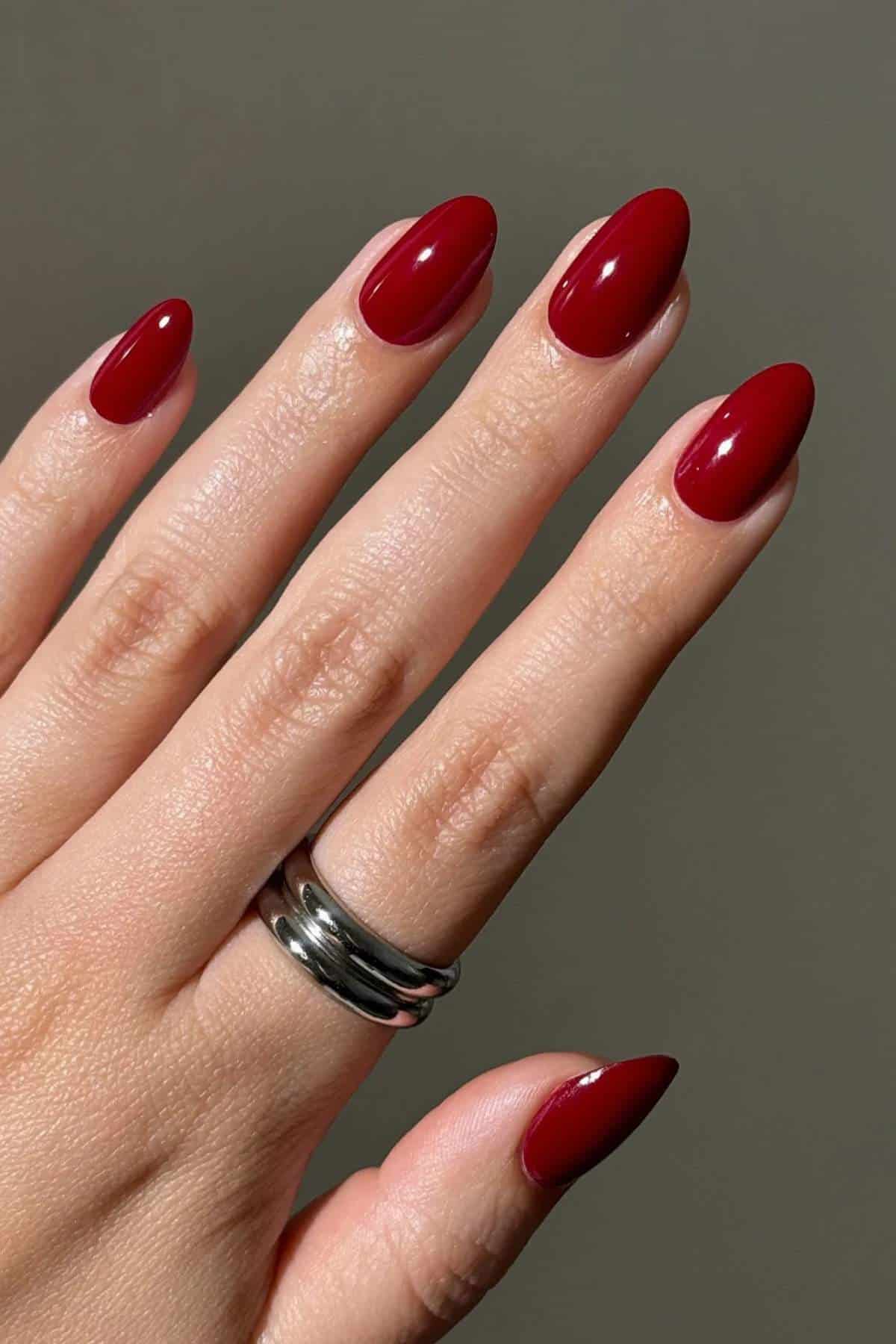 A hand with long, almond-shaped nails painted glossy red, wearing a silver double-band ring on the middle finger.