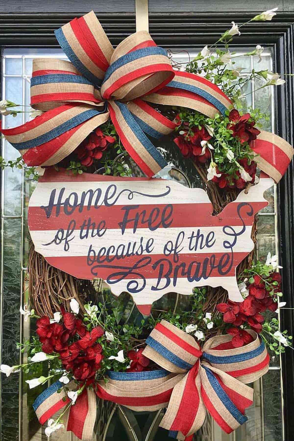 A decorative wreath with red, white, and blue ribbon, red flowers, and a USA-shaped sign that reads, "Home of the Free because of the Brave.