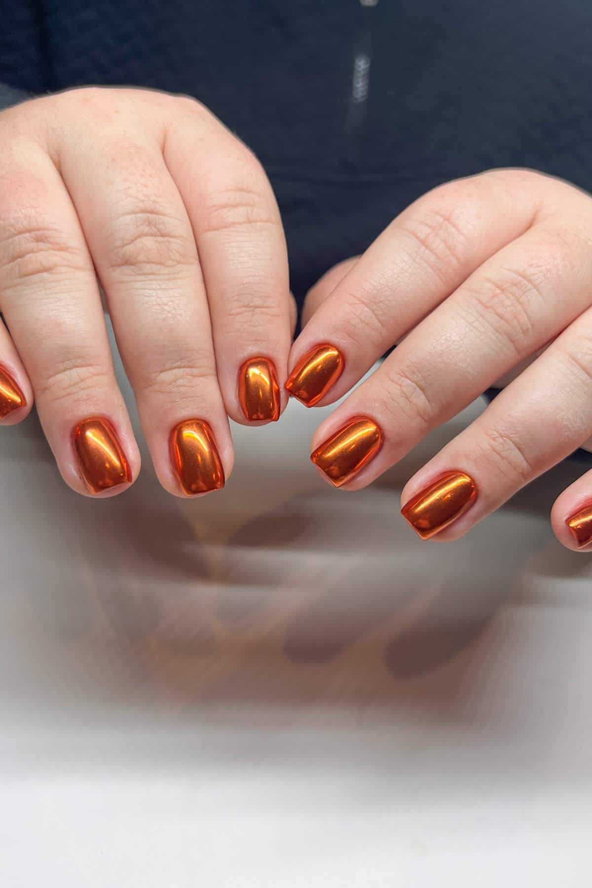 Close-up of two hands with short, square-shaped fingernails painted in a shiny metallic orange polish.