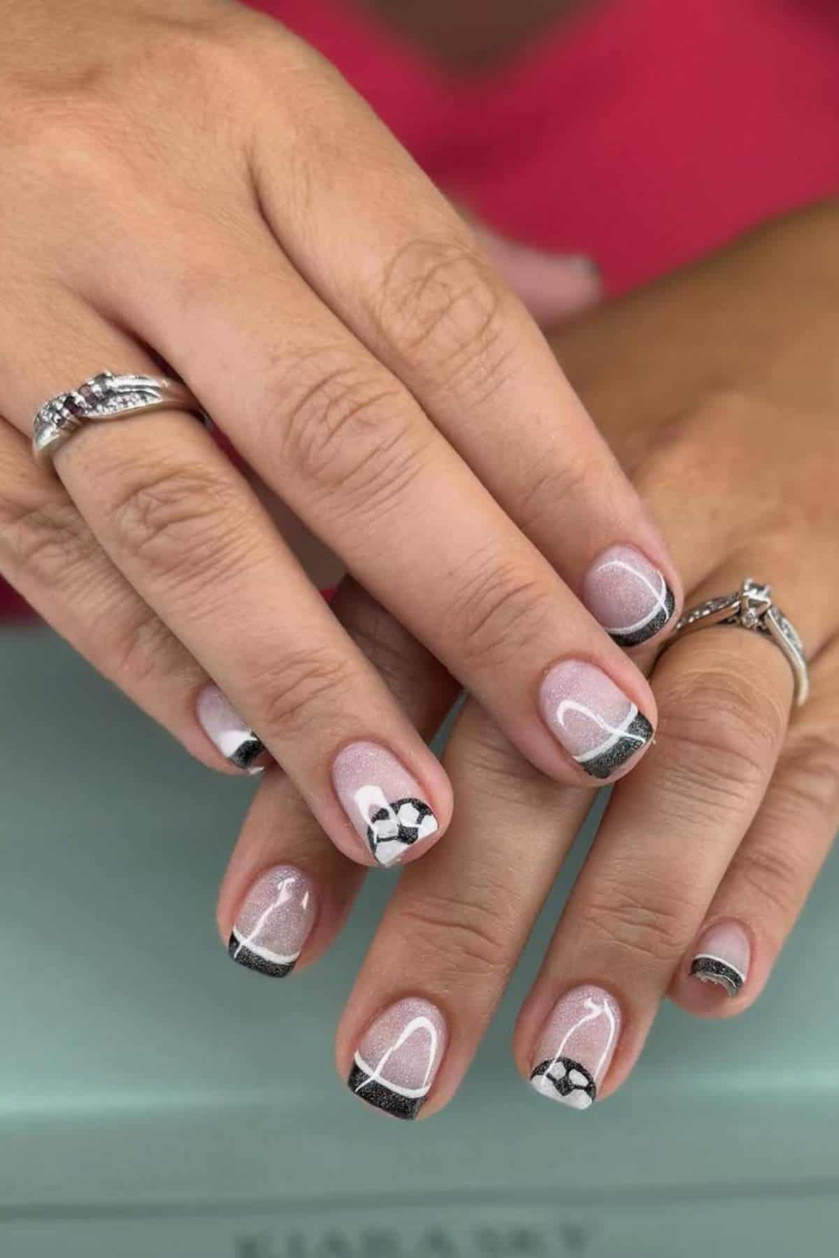Close-up of hands with short, glossy nails featuring black and silver tips, white and black floral designs, and geometric lines; person is wearing silver rings.