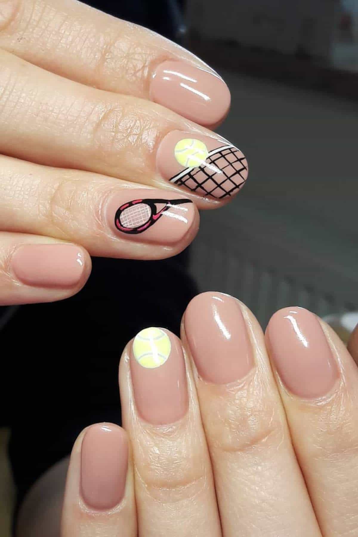 Close-up of hands with nude-colored manicure; two nails feature tennis-themed designs with a racket, net, and yellow tennis balls.