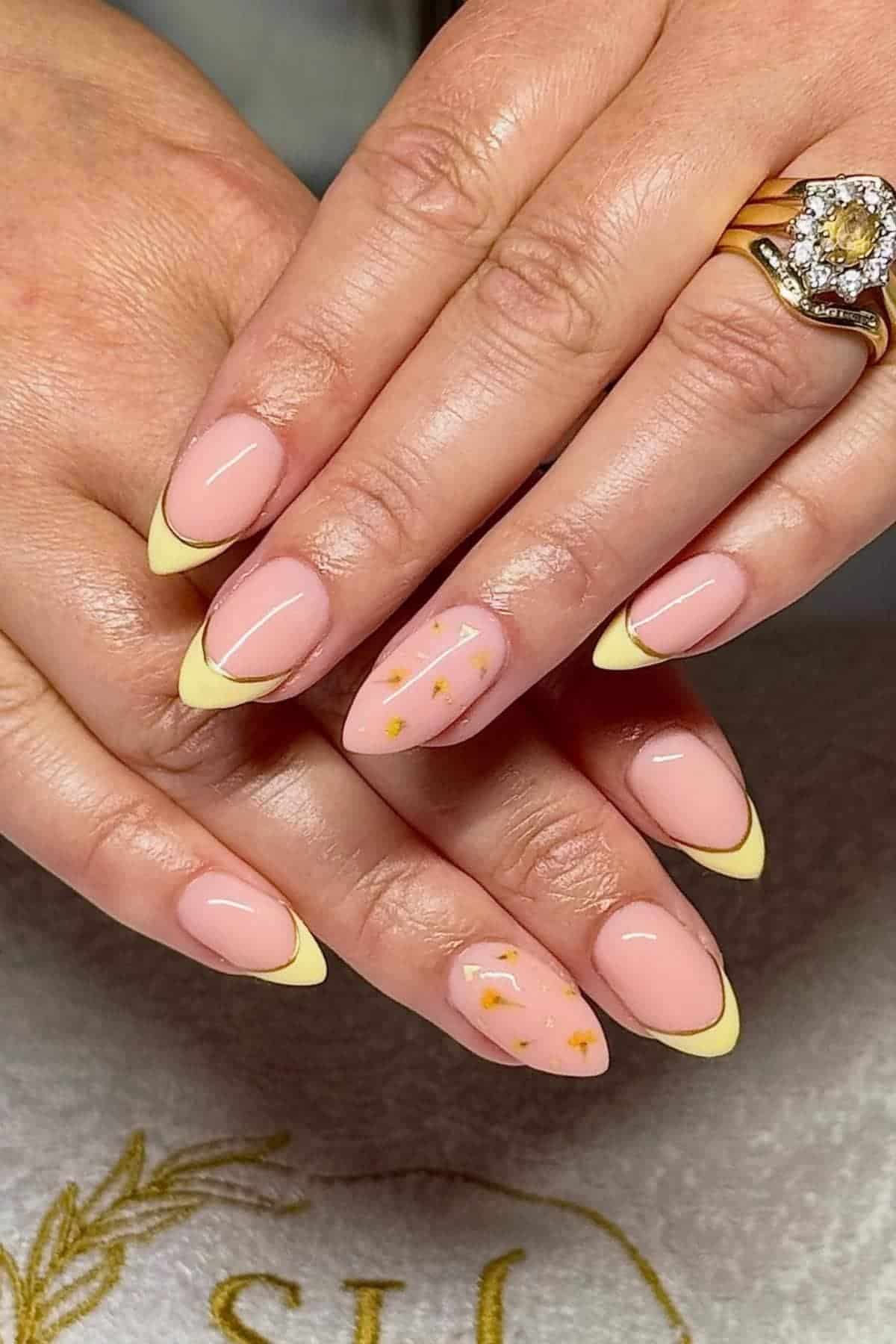 Hands with almond-shaped nails featuring a pale pink base, yellow French tips, and gold foil accents on ring fingers; person is wearing two gold rings.