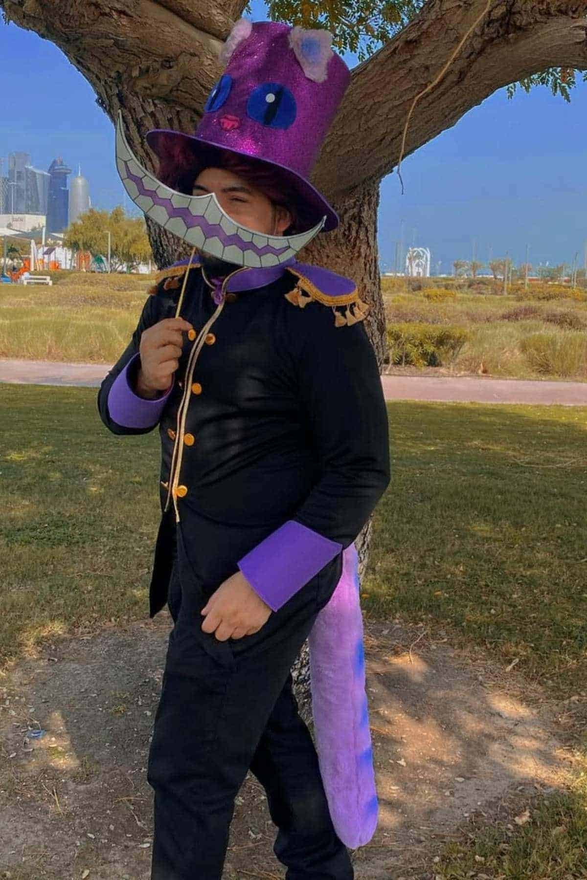 Person dressed in a black and purple costume with a large grin mask and a tall hat, inspired by the Cheshire Cat, standing outdoors near a tree.