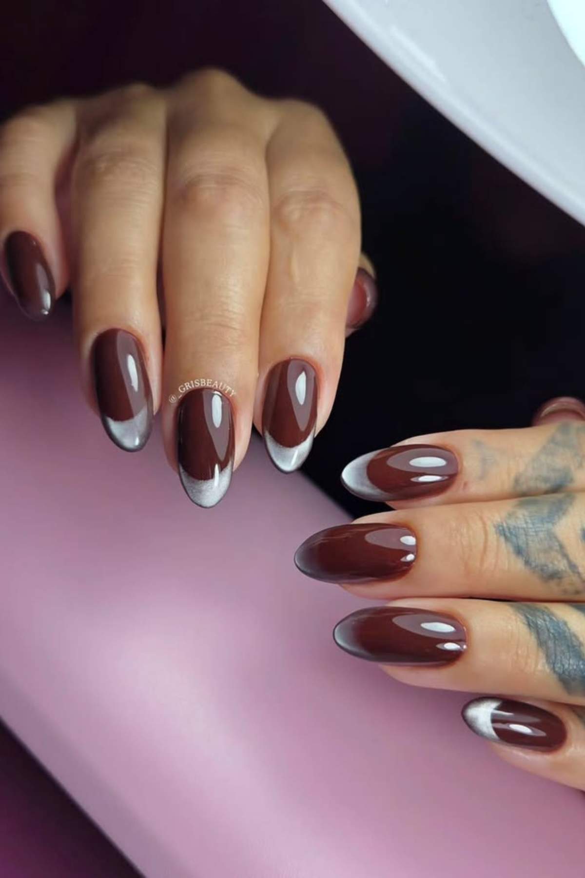 Hands with long oval-shaped fingernails painted in dark brown with silver metallic French tips, posed against a pink surface.