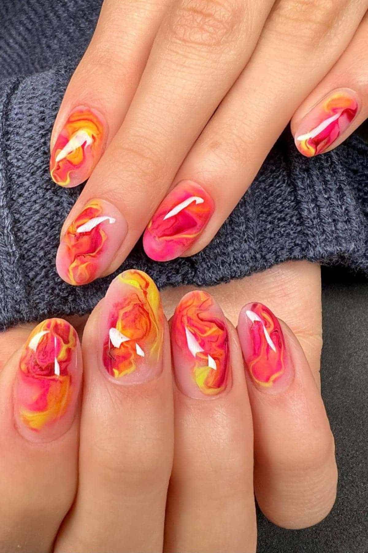 Close-up of hands with almond-shaped nails painted in a glossy, marbled design featuring vibrant shades of orange, yellow, and pink.