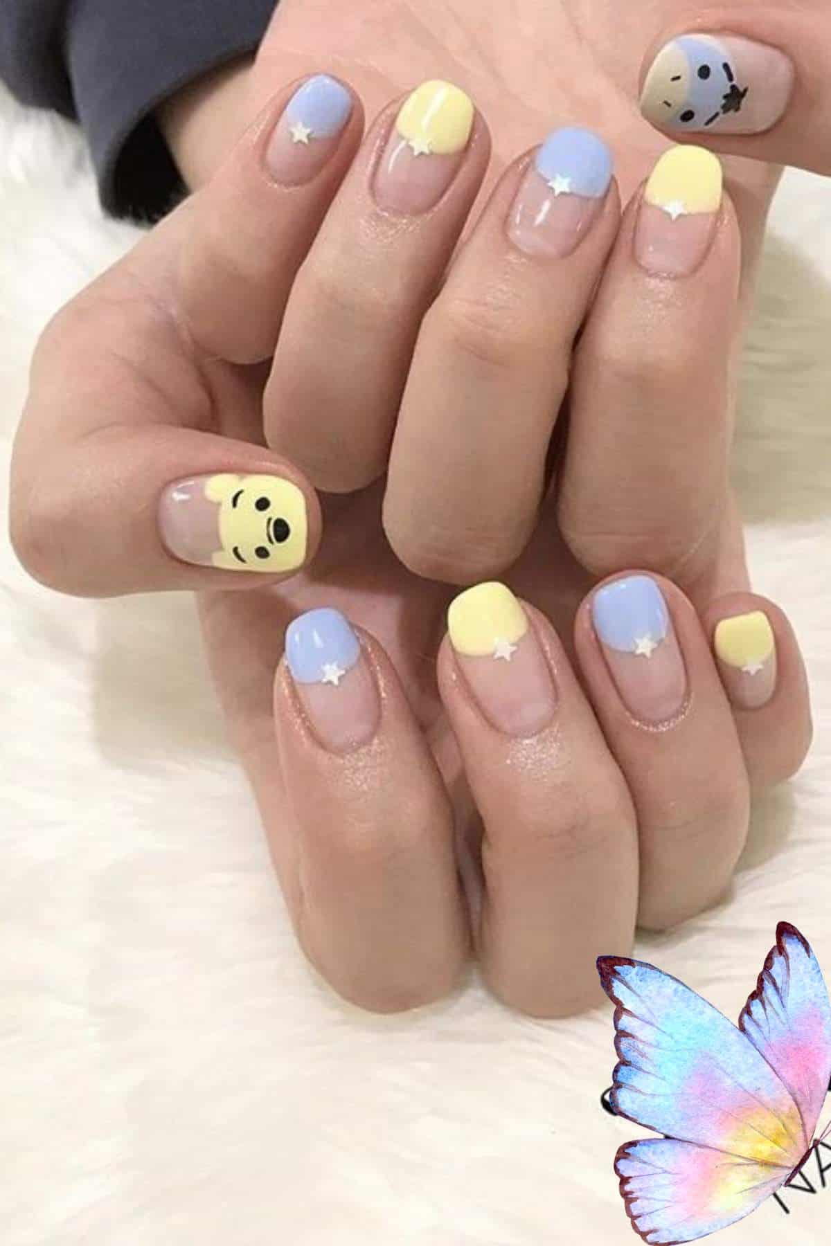 Hands with pastel blue and yellow nail art, featuring small white stars and cartoon faces on two nails, resting on a white surface.