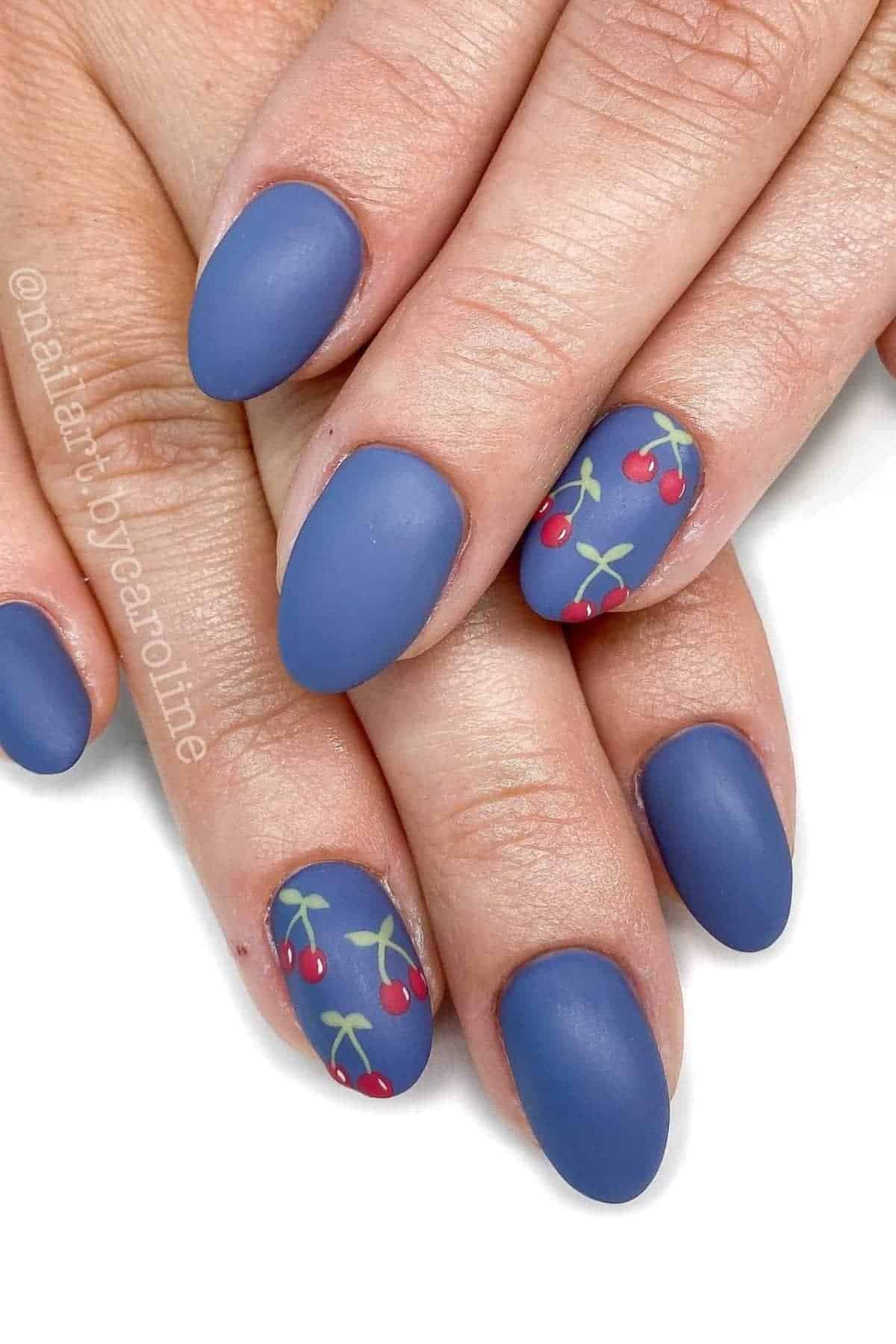 Hands with matte blue almond-shaped nails; some nails feature a small cherry design with green stems and red cherries.