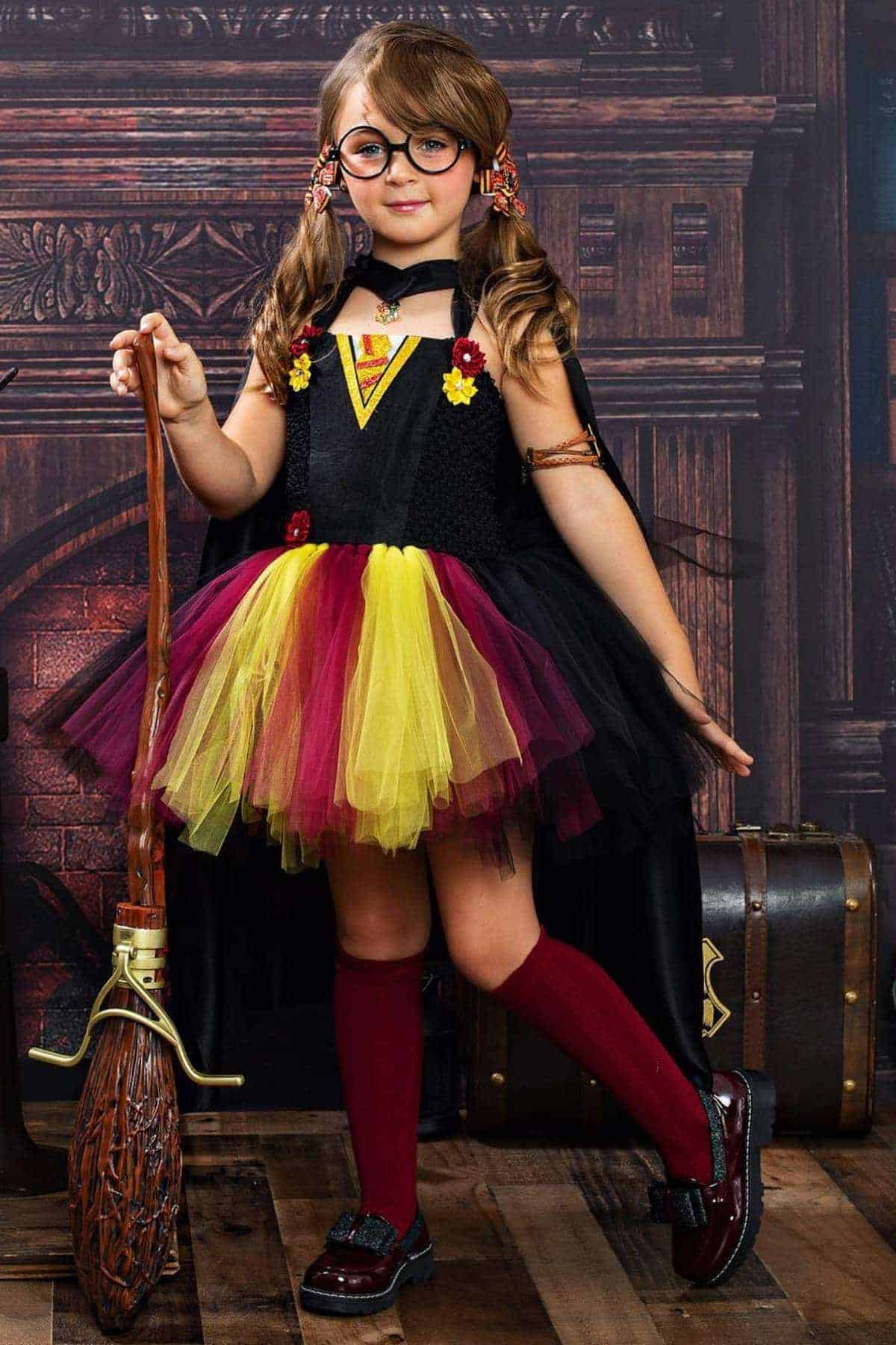 Young girl in a Harry Potter-themed costume with glasses, a cape, and a wand-shaped broom, standing in front of a library backdrop.