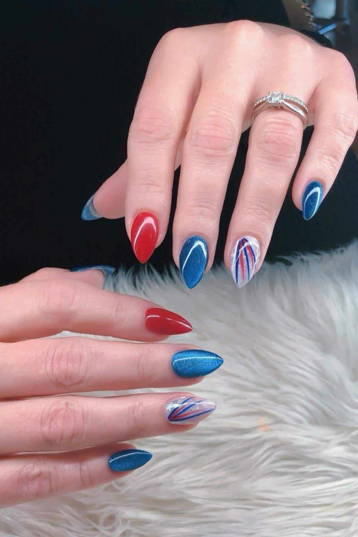 Hands with almond-shaped nails painted in red, blue, and white patterns; some nails have solid colors, others have lines or abstract designs. The person wears a ring on one finger.