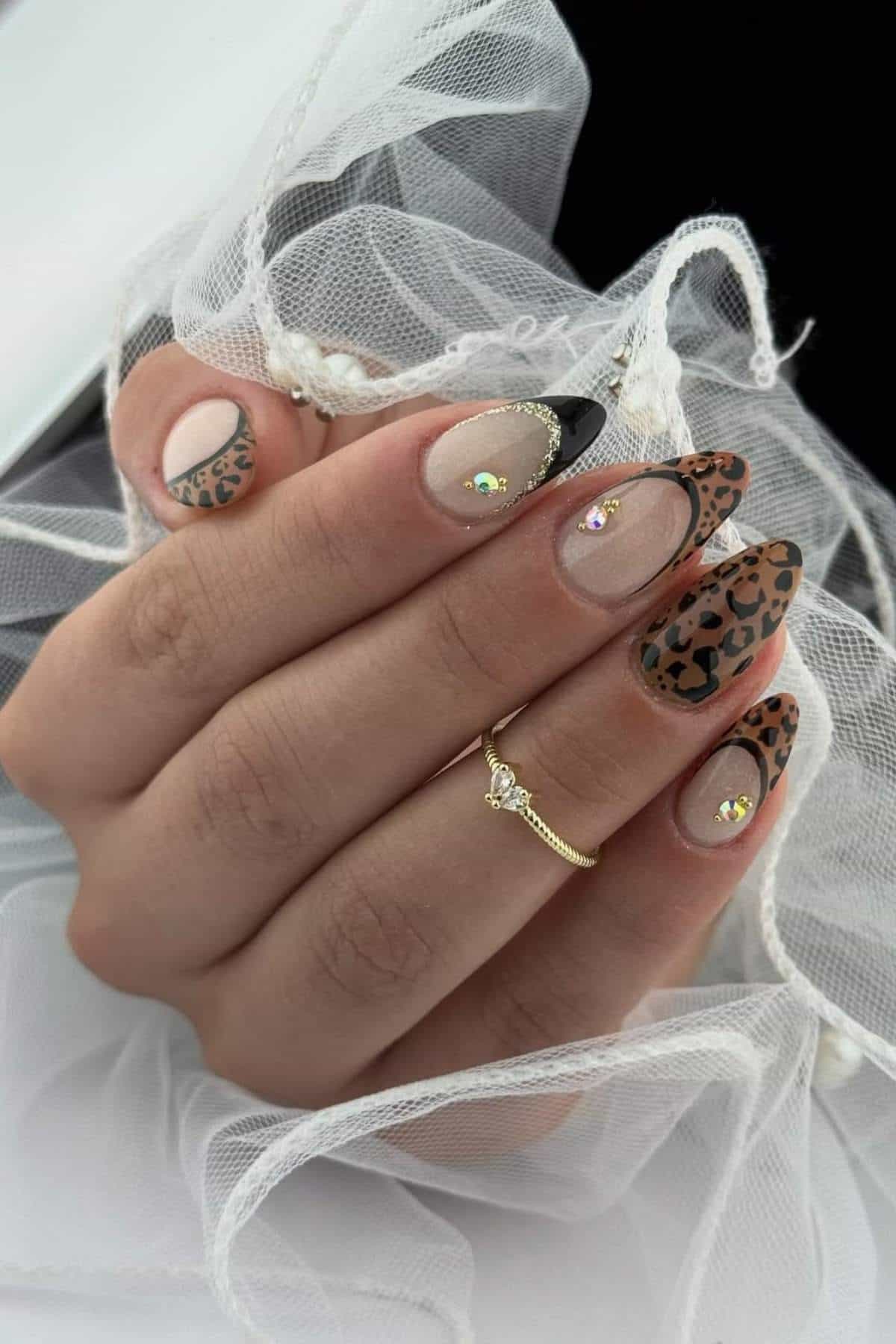 A hand with almond-shaped nails featuring leopard print, nude polish, black tips, and rhinestone accents, holding white tulle fabric.