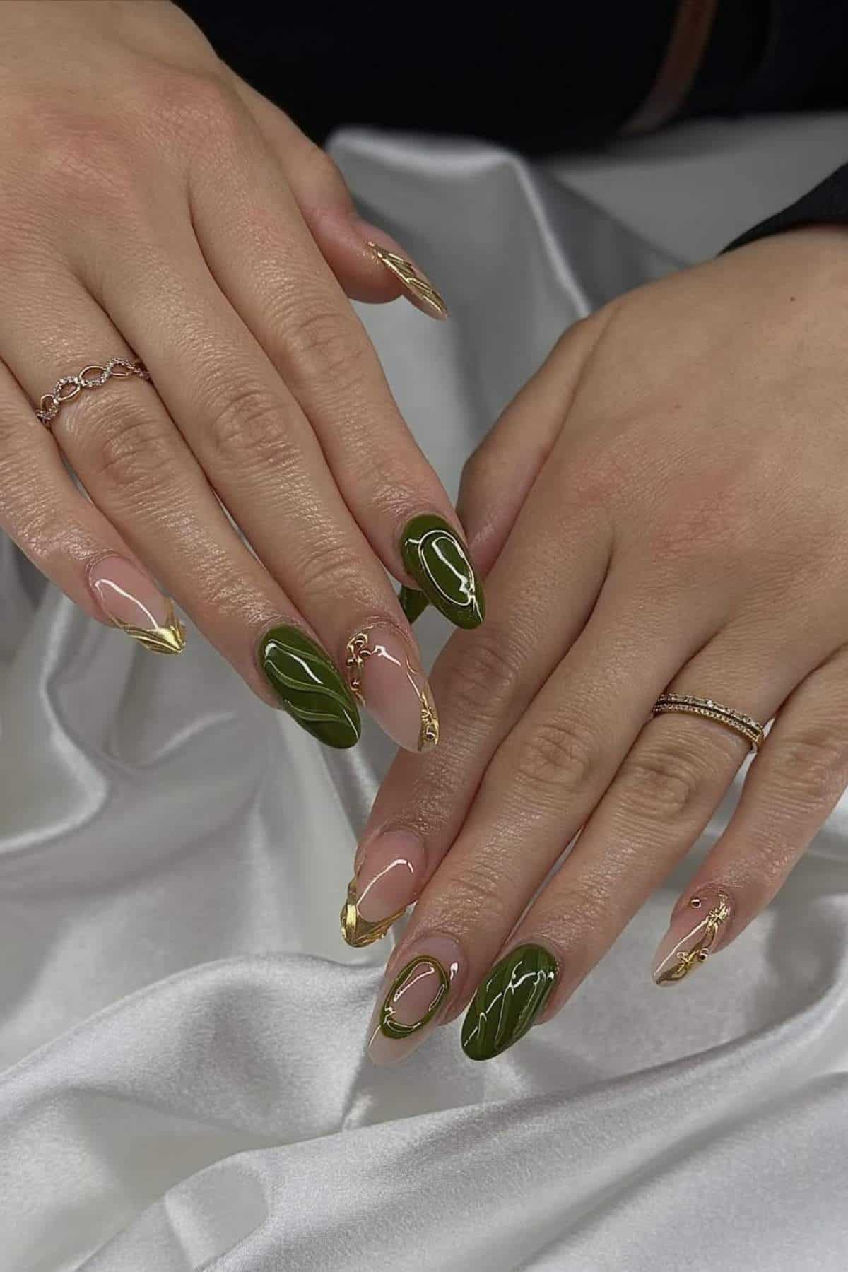 Two hands with long, almond-shaped nails featuring green, gold, and nude nail art designs, resting on a white satin fabric.