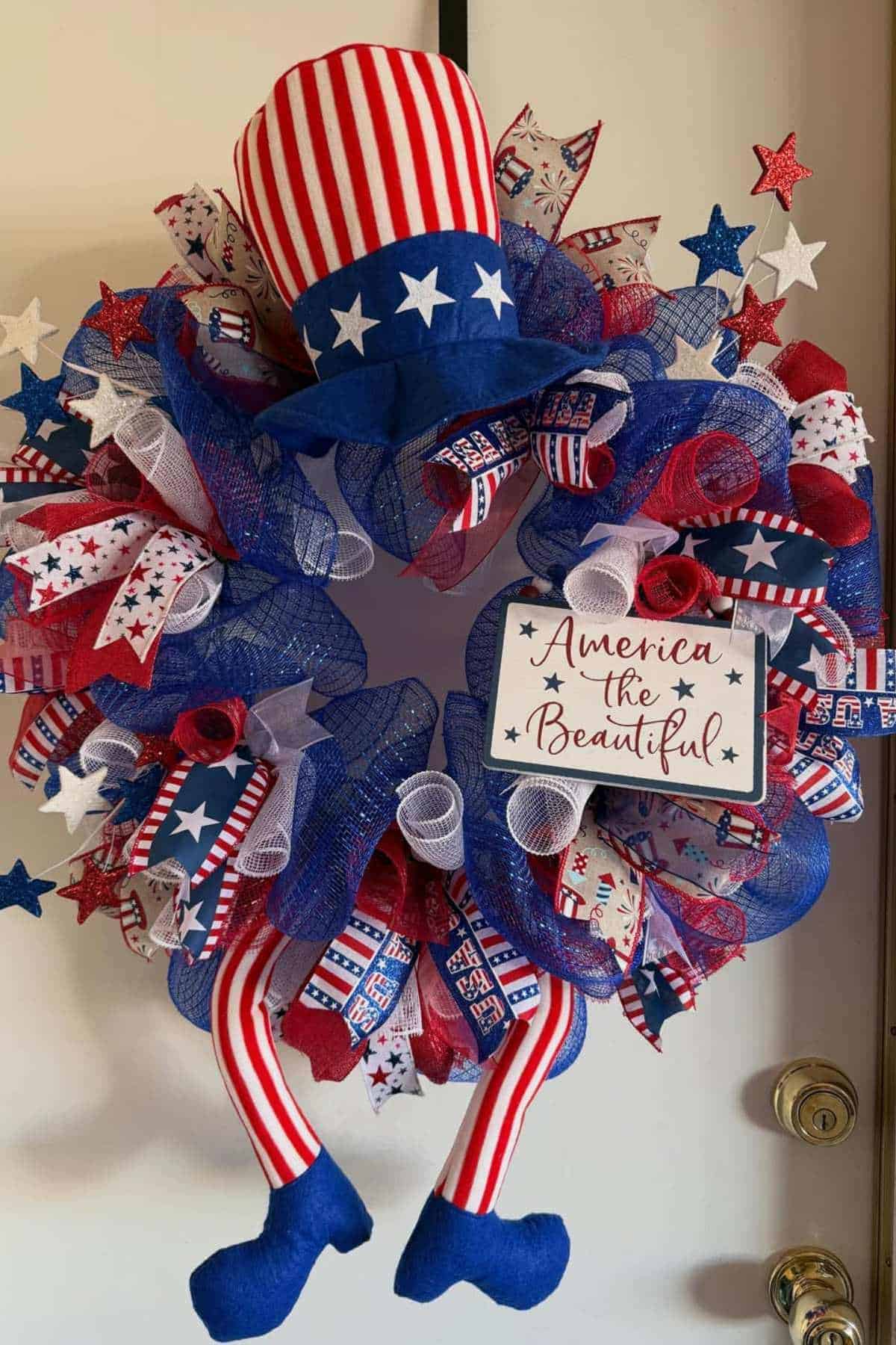 A patriotic wreath decorated with red, white, and blue ribbons, a striped hat, star accents, and a sign that reads "America the Beautiful.