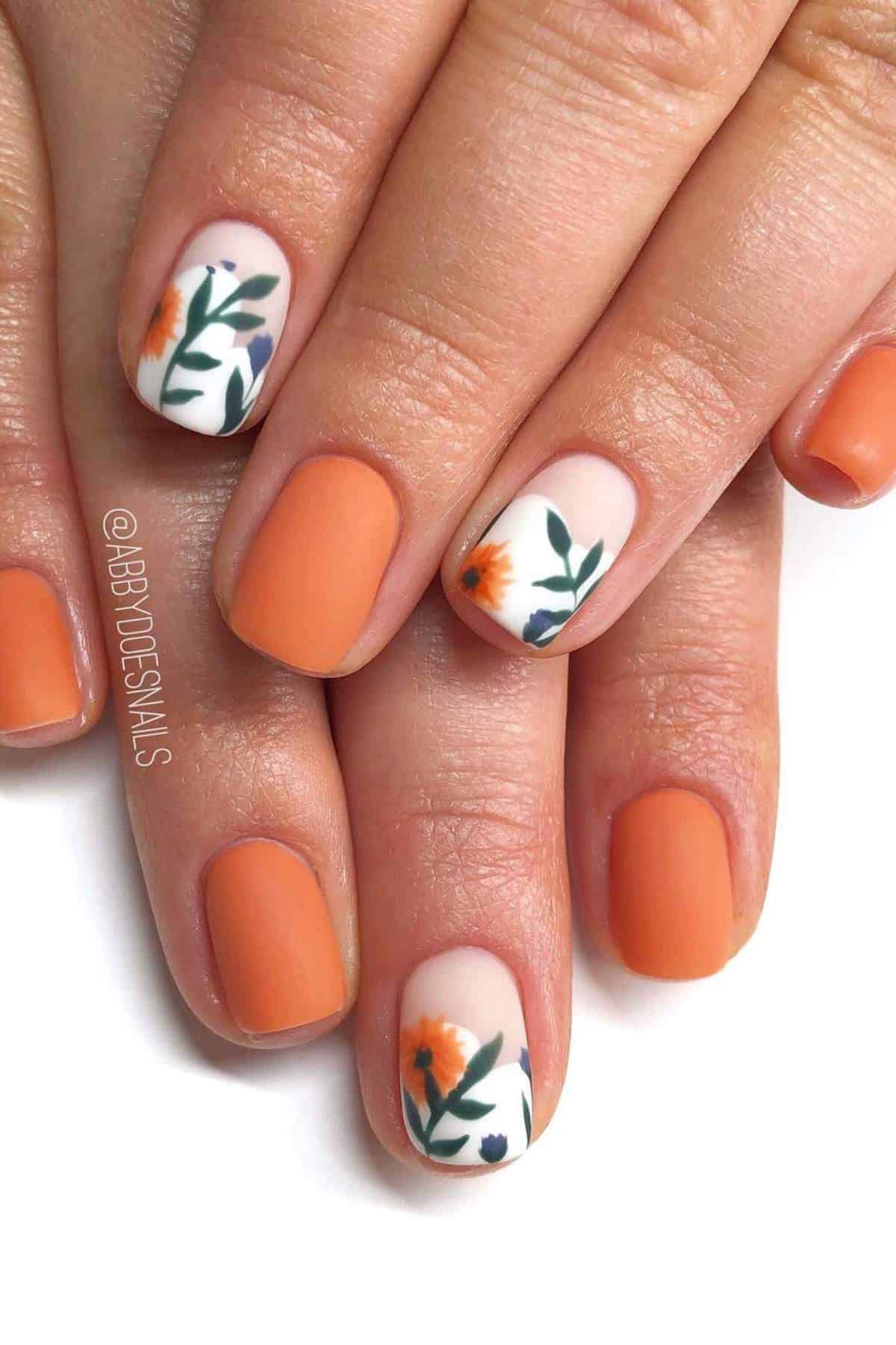 Hands with short, matte orange nails; two accent nails feature a white base with an orange flower and green leaf design.