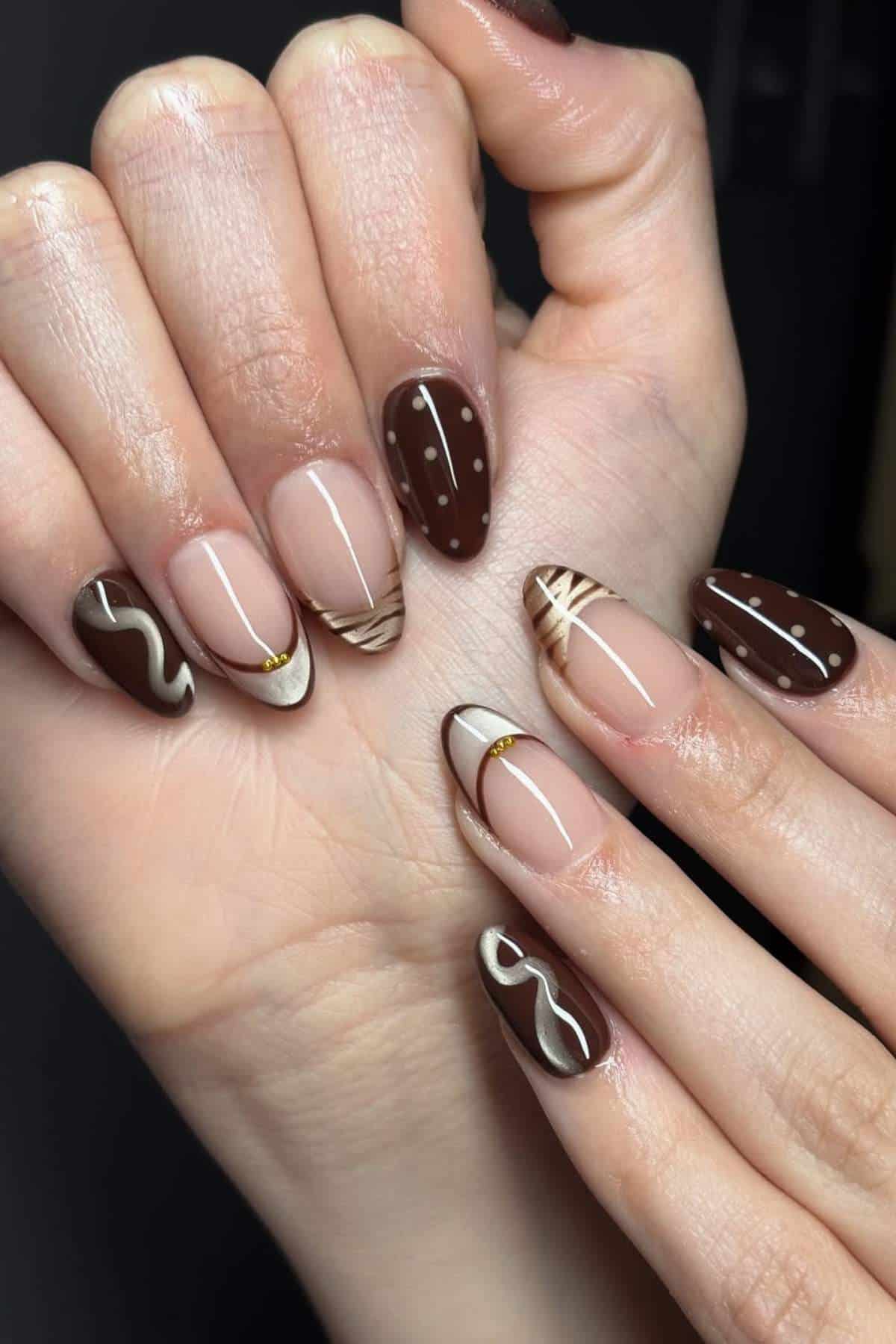 Close-up of hands with almond-shaped nails featuring brown, beige, and gold abstract nail art designs, including dots, swirls, and geometric patterns.