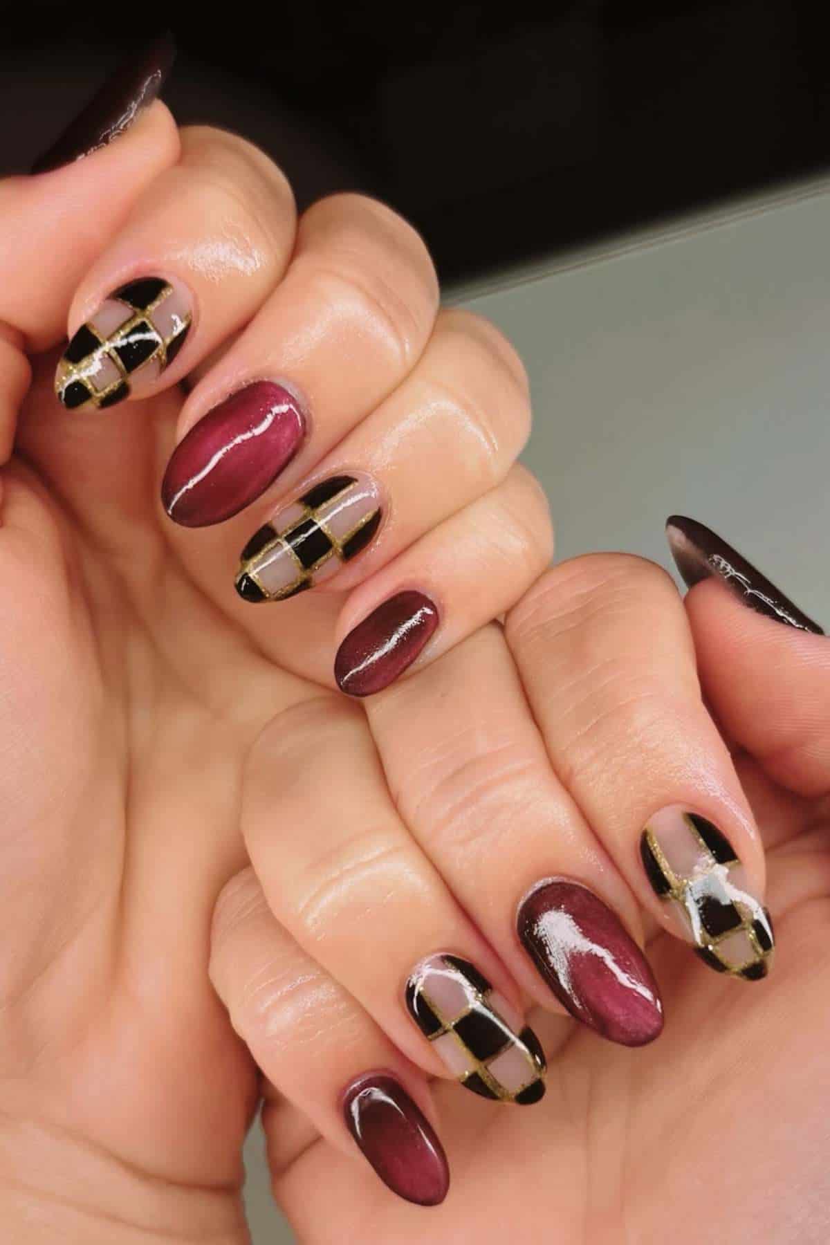 Close-up of hands with manicured nails featuring a burgundy polish and accent nails with a black, gold, and nude geometric harlequin pattern.