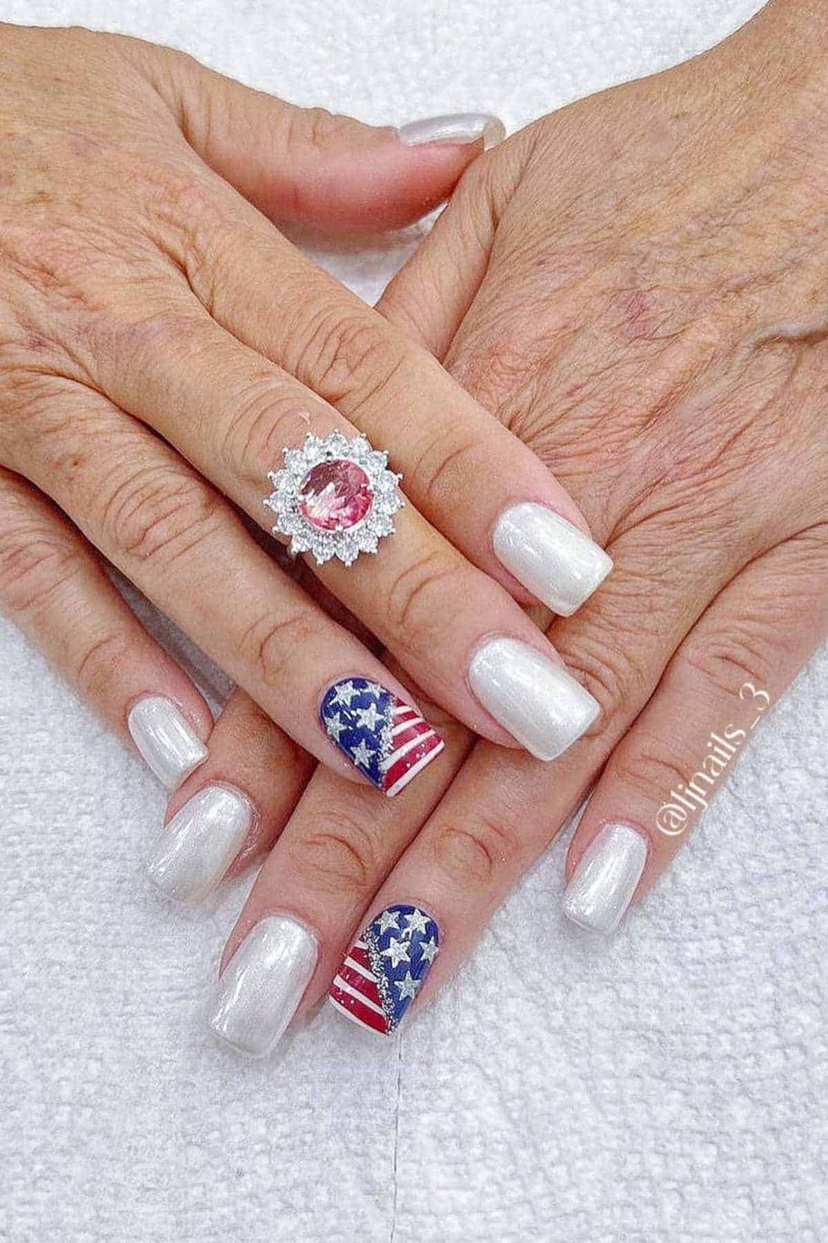 Hands with pearl white nail polish; ring fingers feature an American flag design with stars and stripes. A large ring with a pink stone is on one hand.