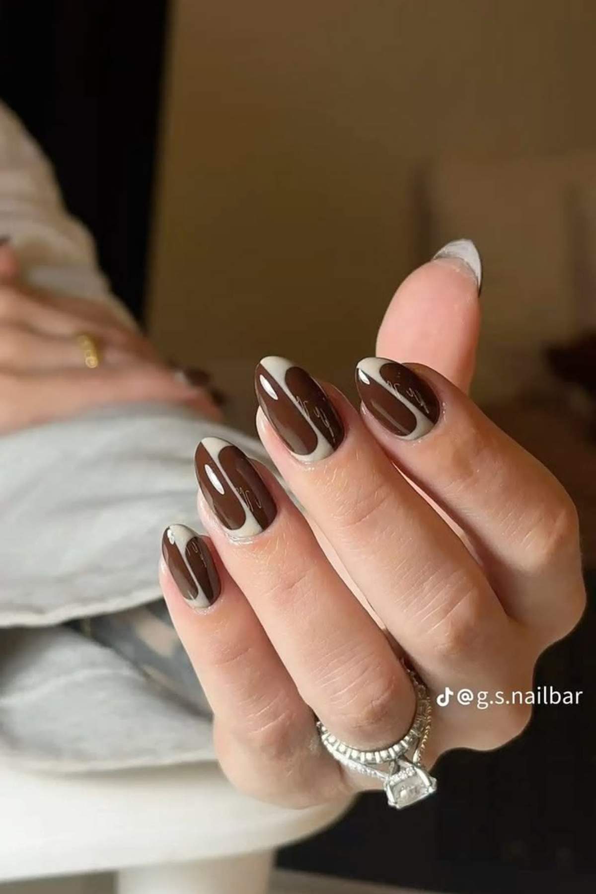 A hand with almond-shaped nails features a brown and beige abstract swirl nail design. The person is wearing silver rings and a diamond ring.