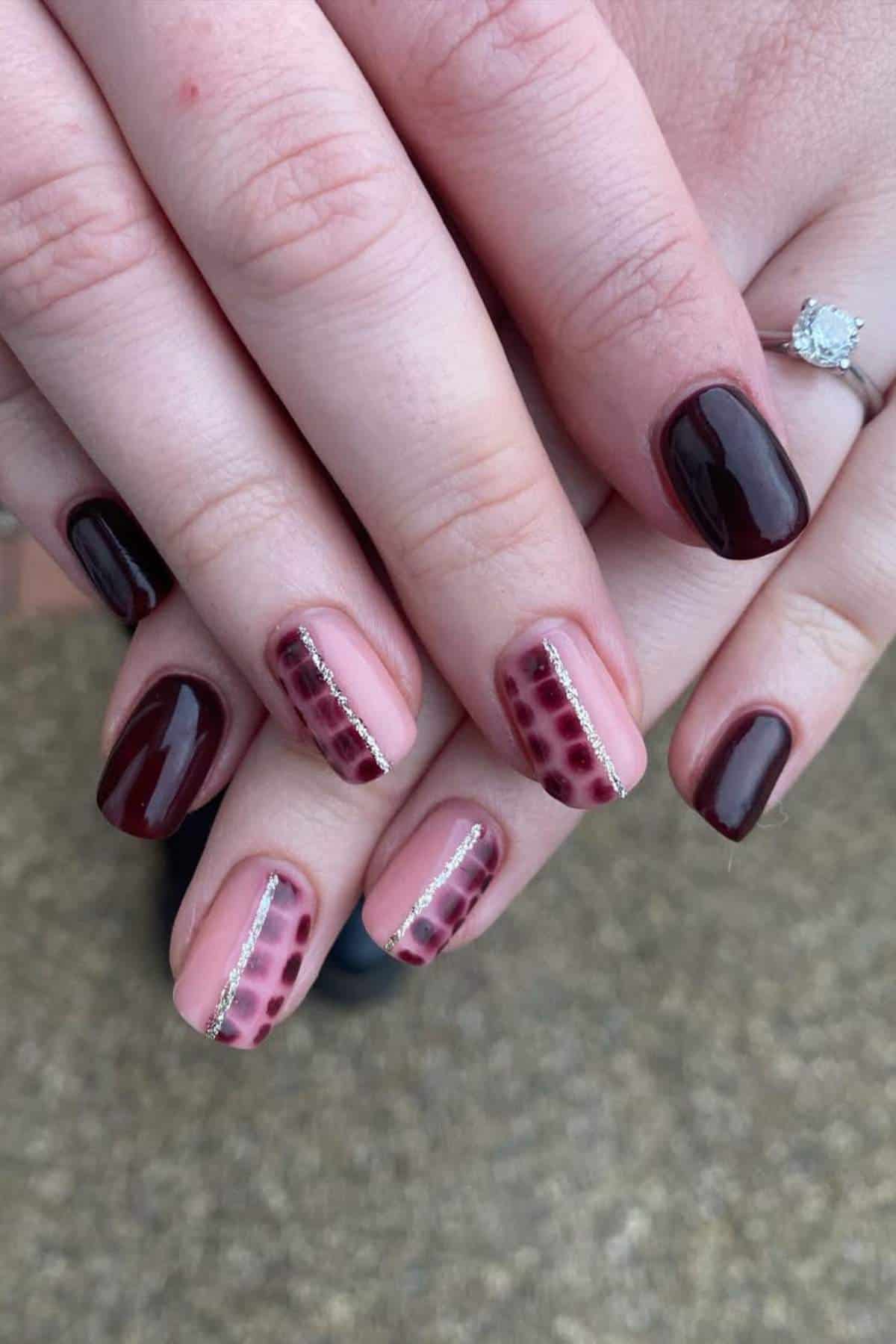 Hands with dark burgundy and nude nails featuring burgundy patterns and silver lines; one hand has a diamond ring on the ring finger.