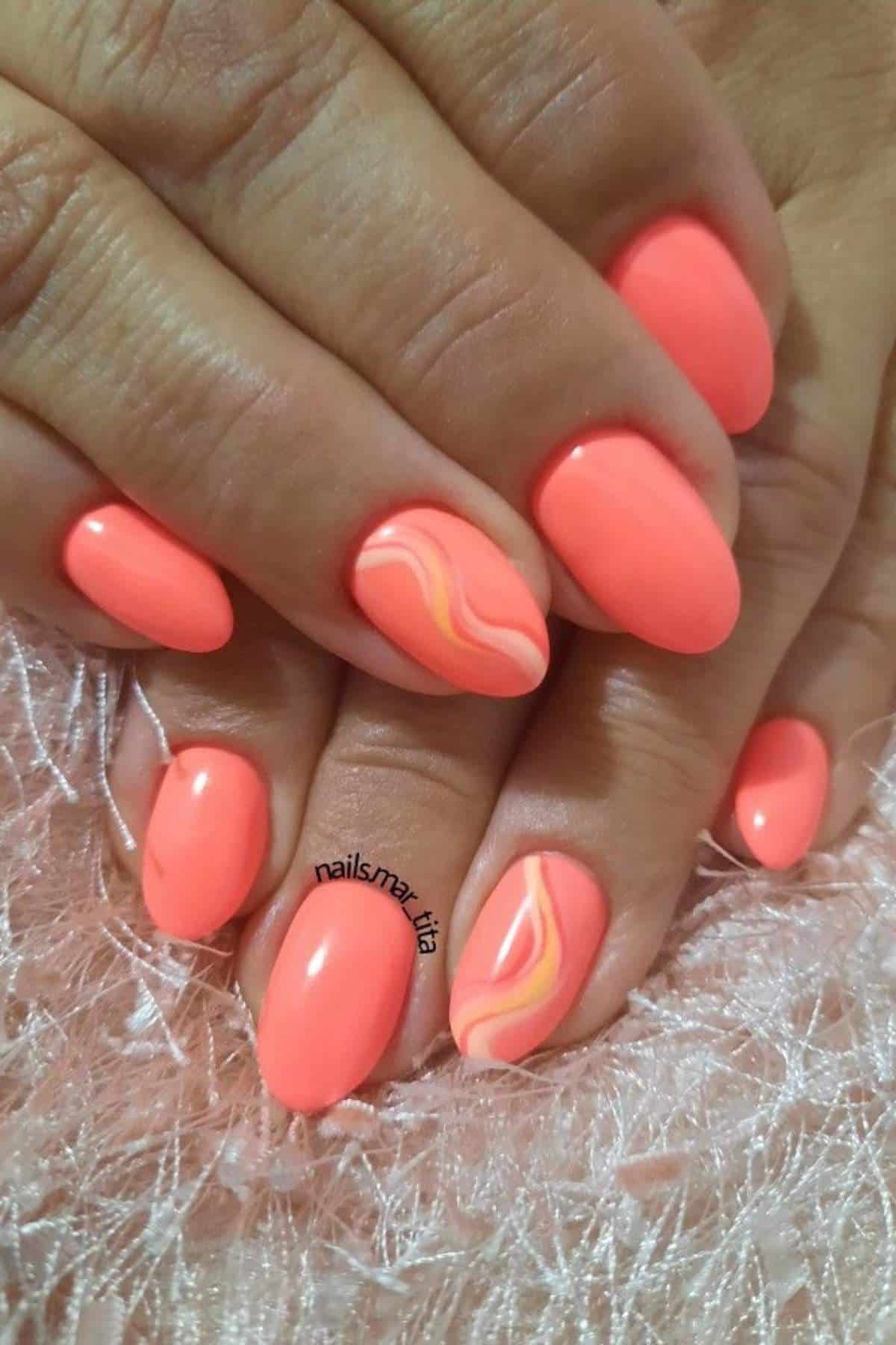Close-up of hands with oval-shaped, bright coral-colored nails; two accent nails feature a wavy marble design in orange, yellow, and white.