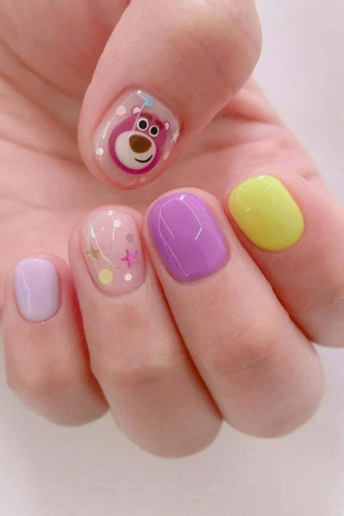 A hand with short nails painted in pastel shades of purple, yellow, and pink, with one nail featuring a bear face and another with small colorful star and dot designs.