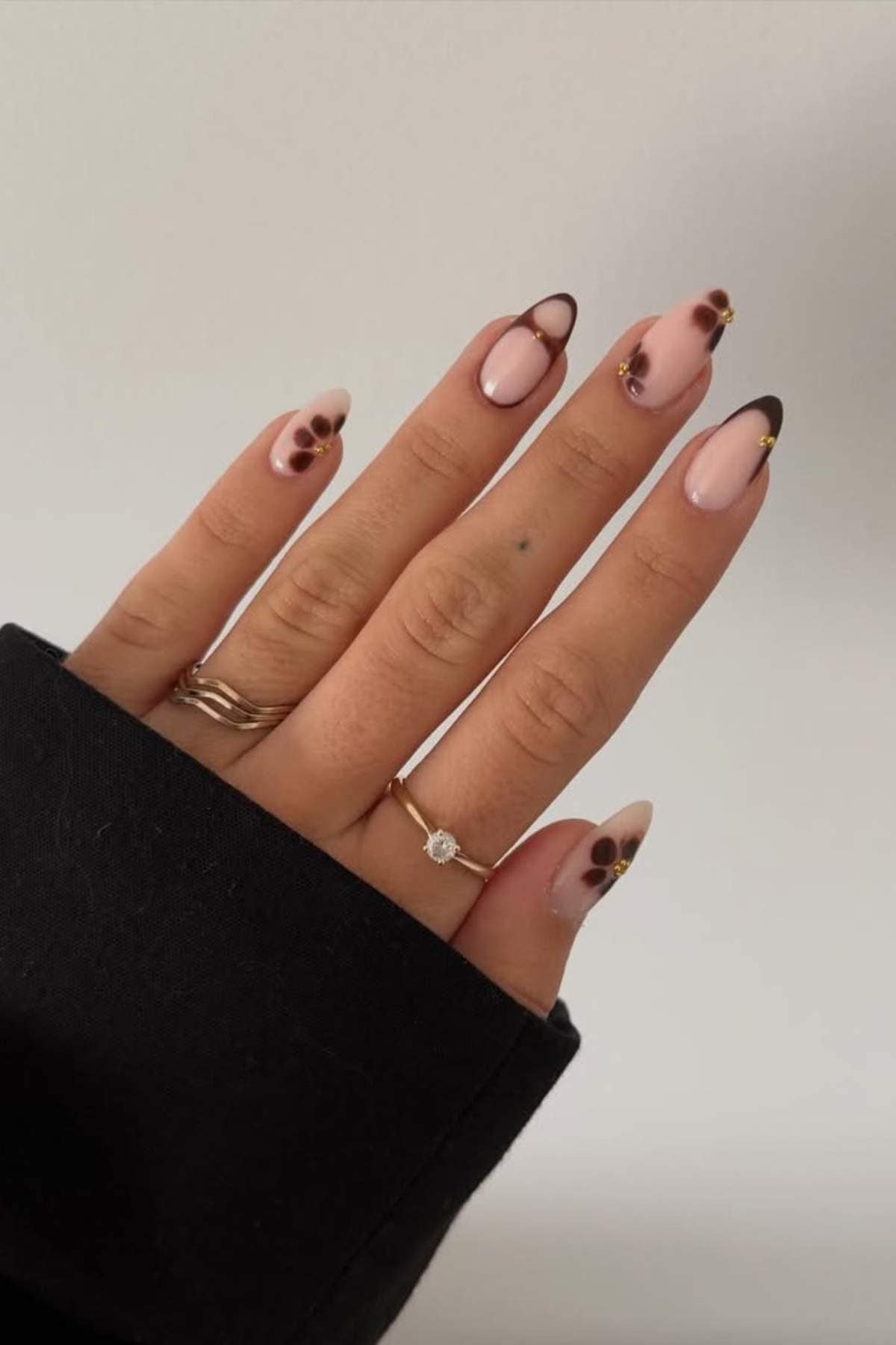 A hand with almond-shaped nails featuring a light pink base, dark brown French tips, gold accents, and small brown dot designs, wearing two gold rings, one with a diamond.