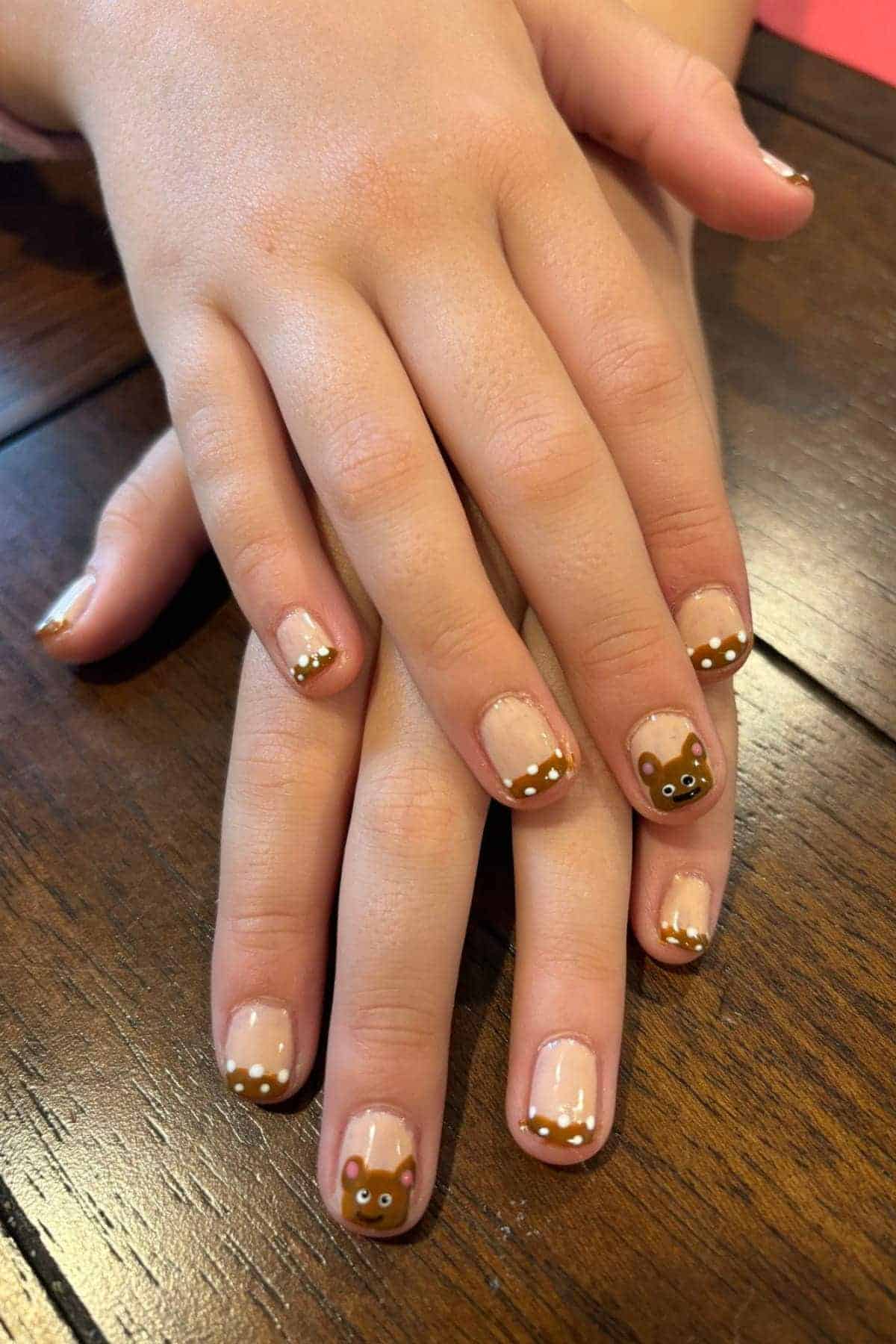 Hands with a manicure featuring bear faces on the thumbnails and brown French tips with white dots on the other nails, resting on a wooden surface.