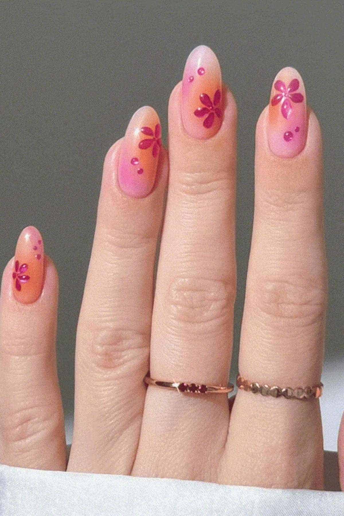 A hand with almond-shaped nails painted in a pink gradient, decorated with red flower designs and small gems, wearing two gold rings.