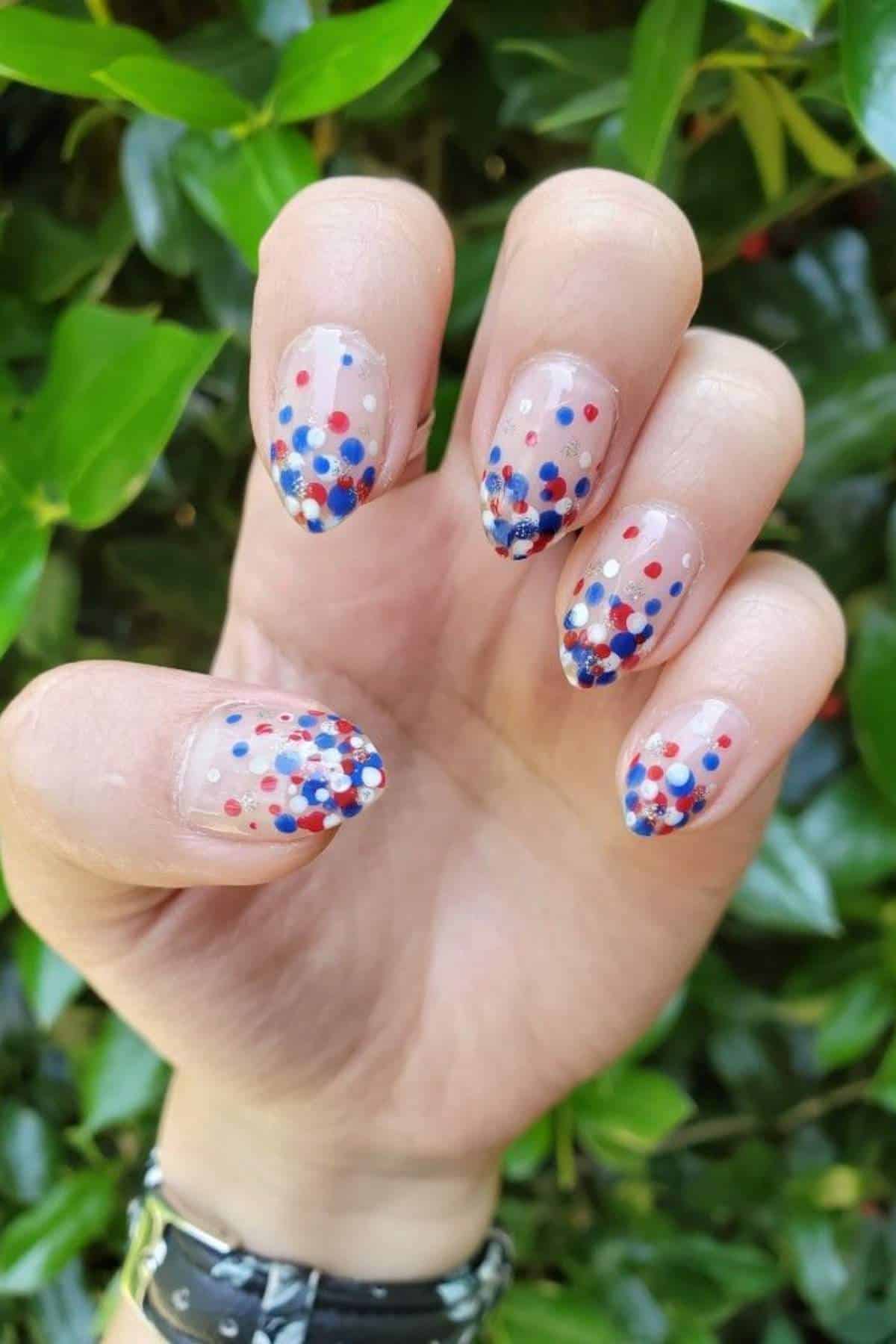 A hand with short, almond-shaped nails featuring clear polish and red, blue, and white confetti dots, held against a background of green leaves.
