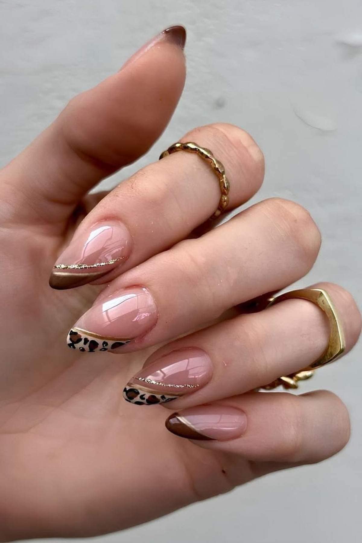 A hand with long almond-shaped nails featuring a brown French tip design, gold accents, and leopard print details; the person is wearing gold rings.