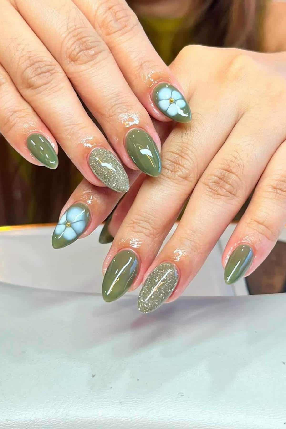 Hands with almond-shaped nails painted in olive green, featuring accent nails with white flower designs and glitter details.