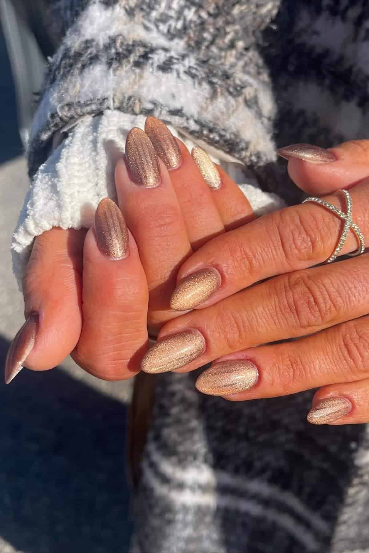 Close-up of hands with long, almond-shaped nails painted in shimmering gold and bronze polish, with one hand wearing a silver ring and a textured sweater sleeve visible.