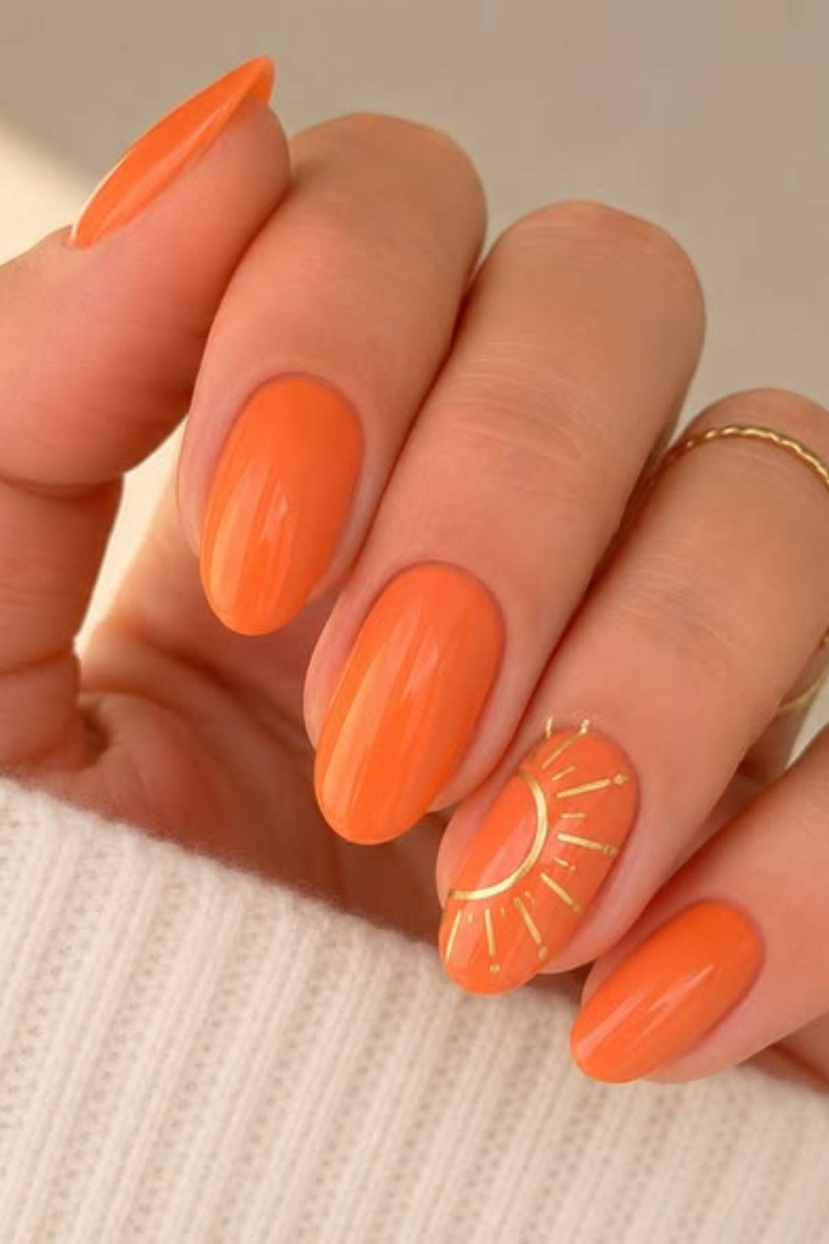 A hand with long, oval-shaped orange nails; the ring fingernail features a sun design in gold.