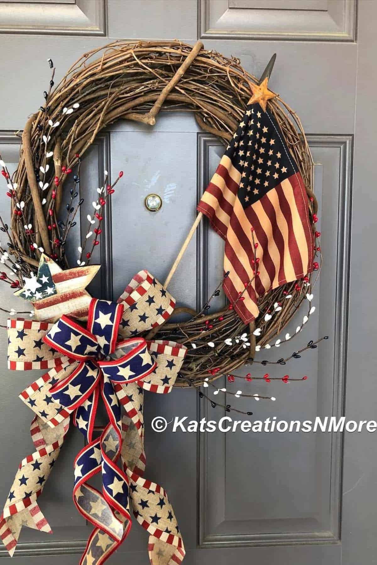 A grapevine wreath decorated with a fabric American flag, star ornaments, berry sprigs, and a large patriotic bow hangs on a gray door.