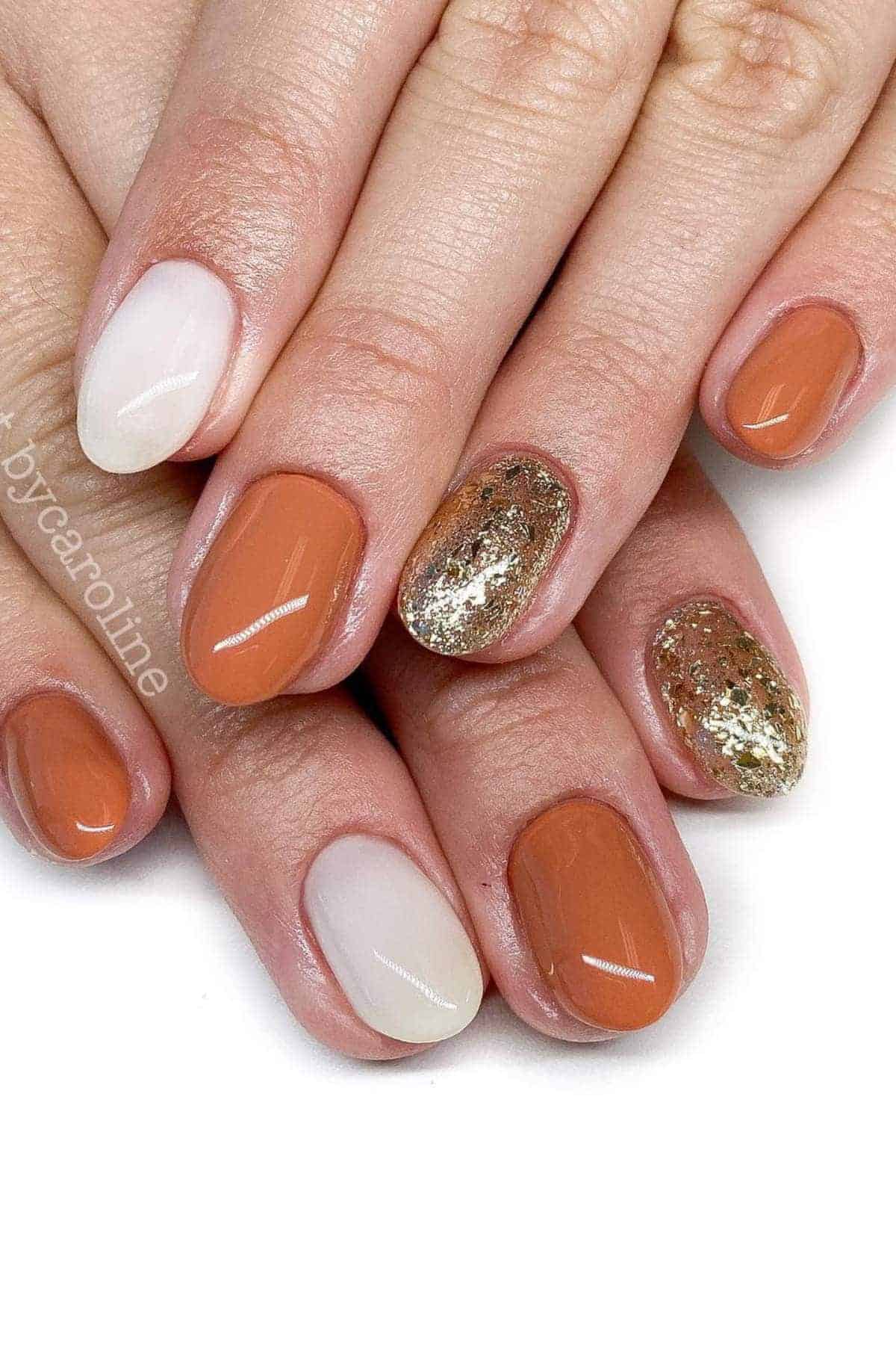 A hand with oval-shaped nails featuring a mix of solid white, burnt orange, and gold glitter polish.