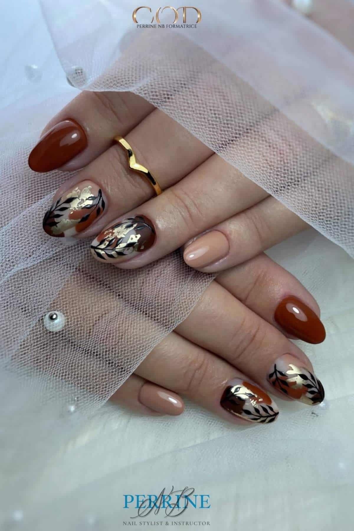 A hand with brown and beige autumn-themed nail art featuring leaf and metallic designs, displayed against a sheer white fabric.